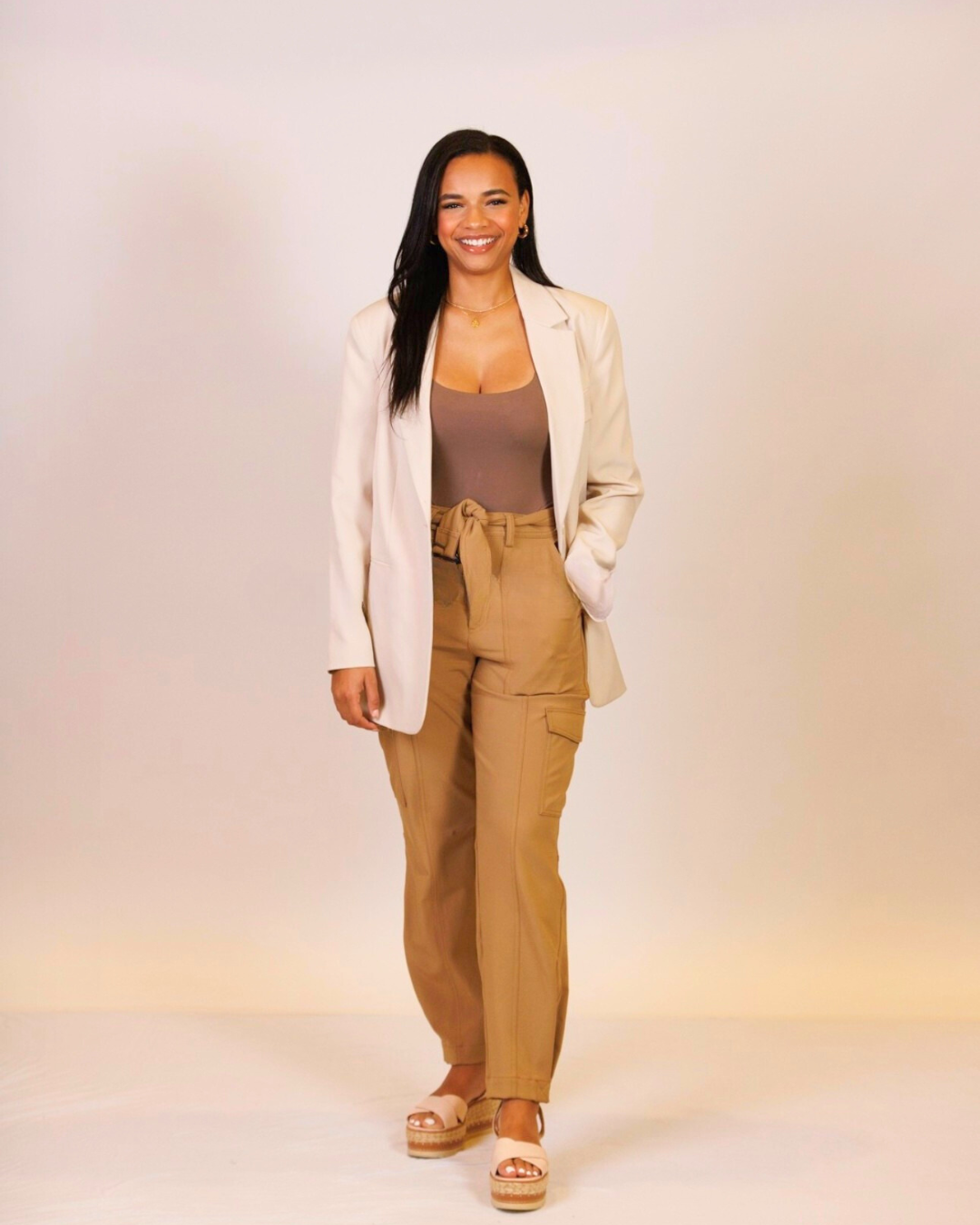 A woman wearing a white blazer and khaki pants is standing in front of a white wall.