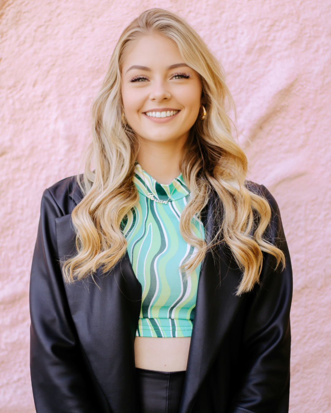A woman wearing a black jacket and a green crop top is smiling.