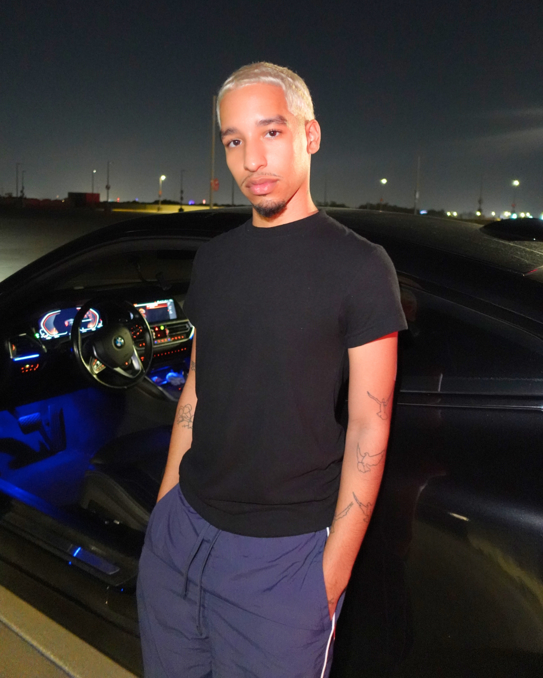 A man in a black shirt and blue shorts is standing in front of a black car.