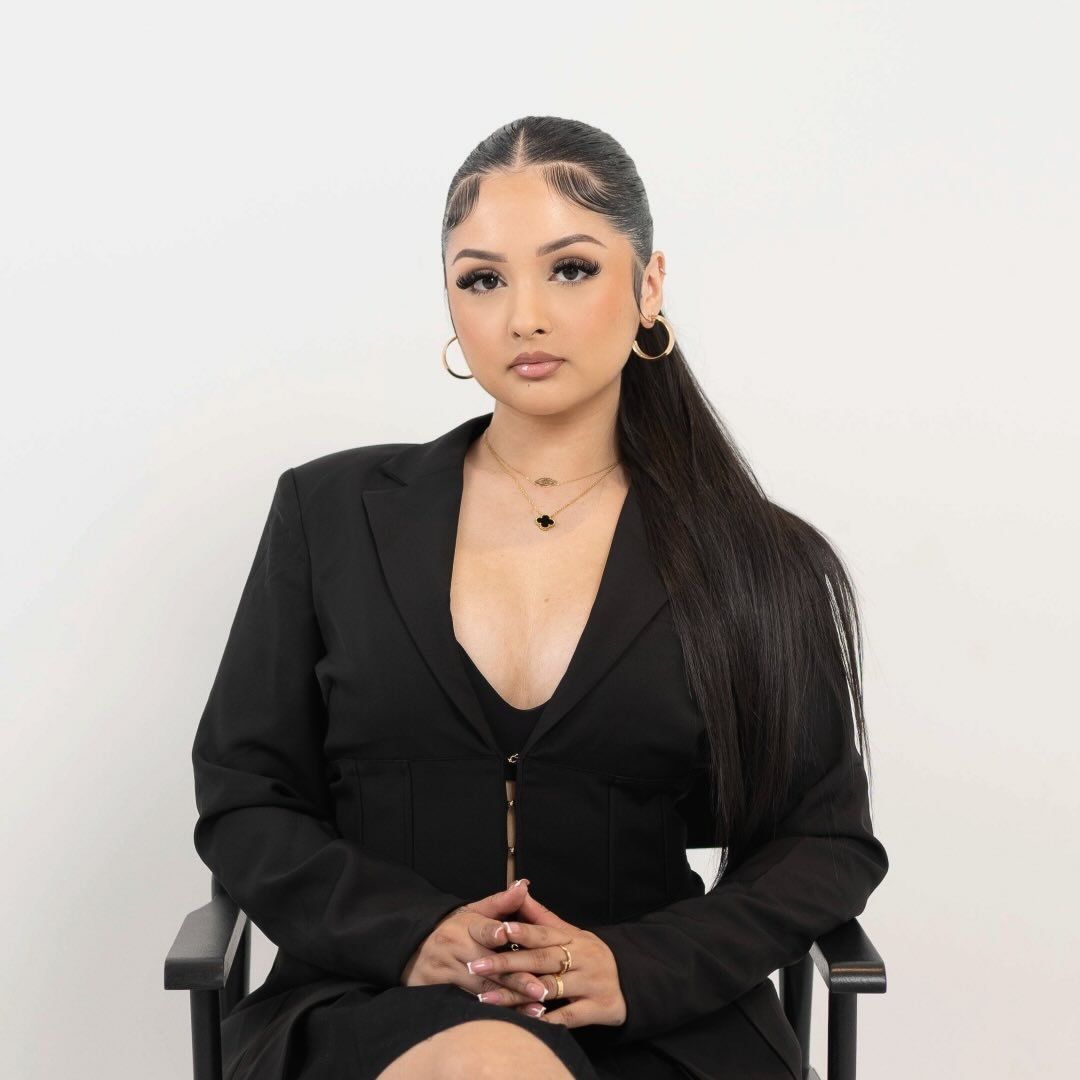 Woman in a black blazer and ponytail, seated and looking forward, hands clasped.