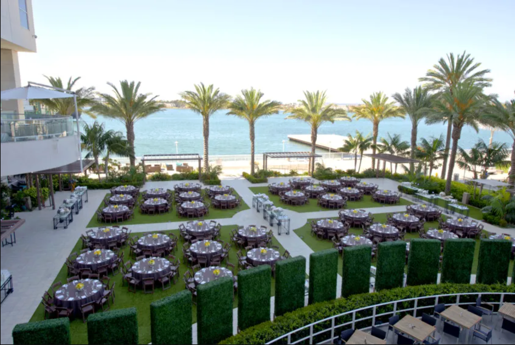 An outdoor seaside venue set up for an event with many round tables and chairs on a lawn surrounded by palm trees.
