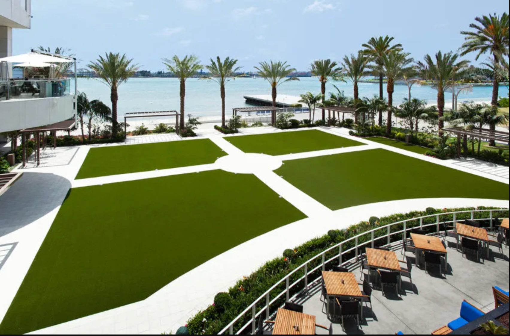 An outdoor patio area with tables overlooking a green lawn divided into four sections, palm trees, and a distant bay.
