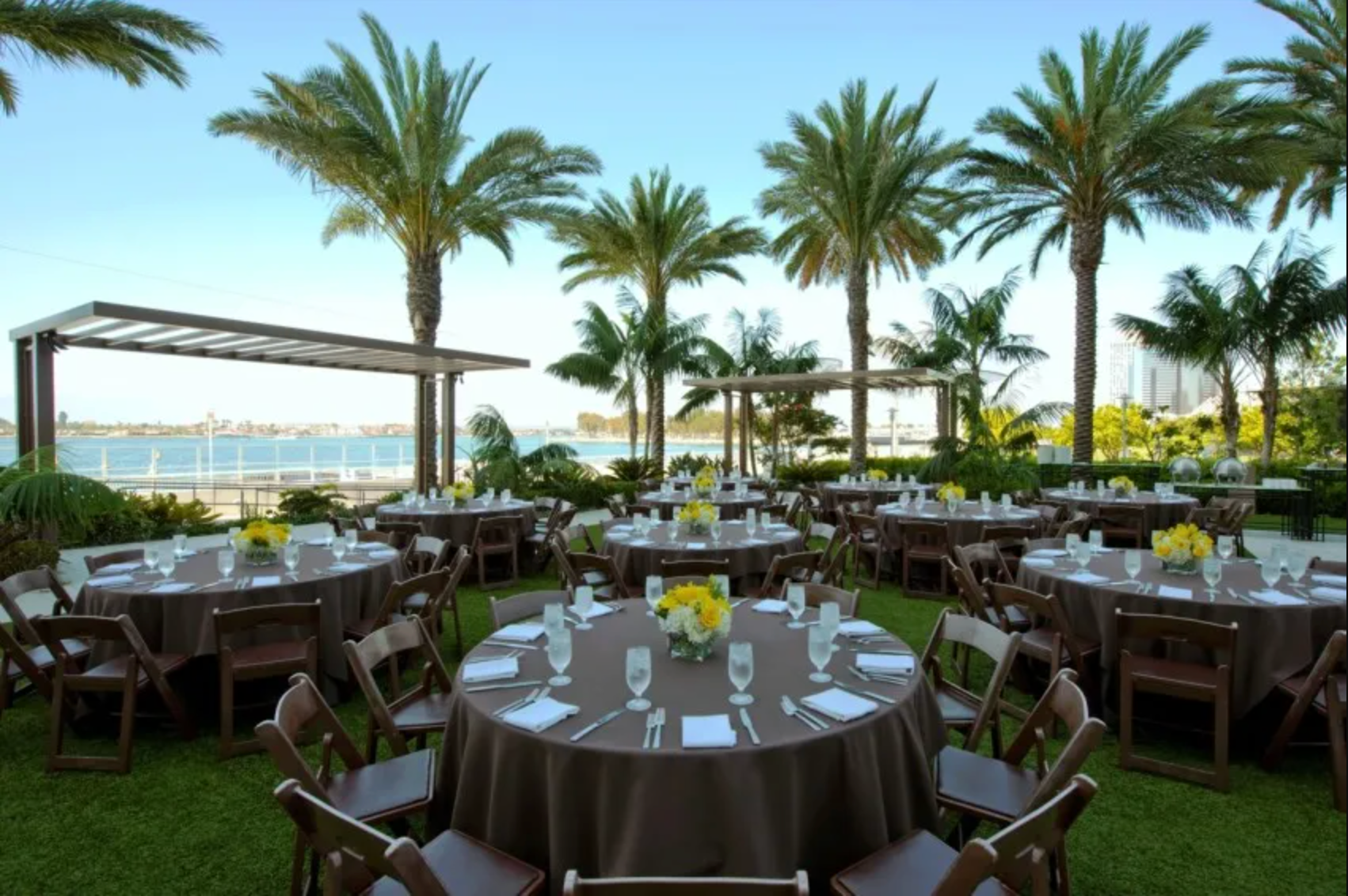 Outdoor venue with round tables set for a meal, featuring brown tablecloths, floral centerpieces, and palm trees.