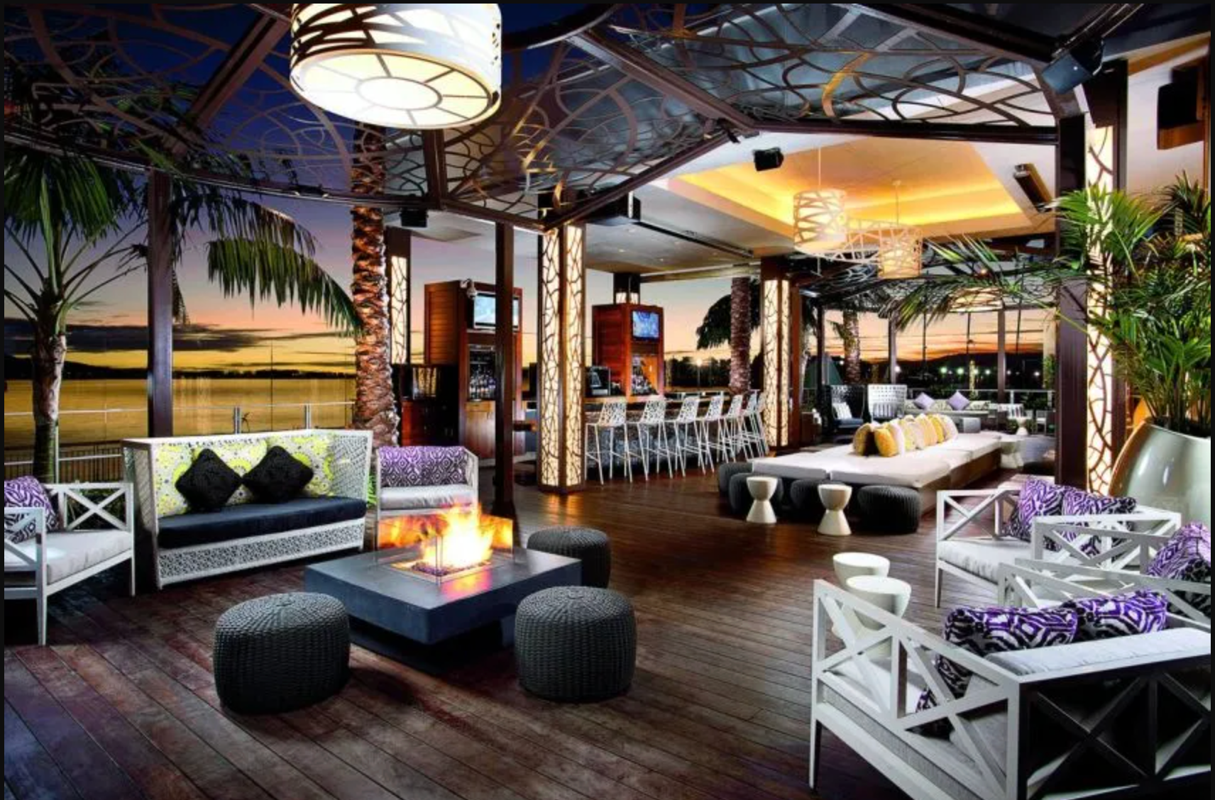 An outdoor lounge with white furniture, fire pit, and bar overlooking a body of water at dusk with warm ambient lighting.