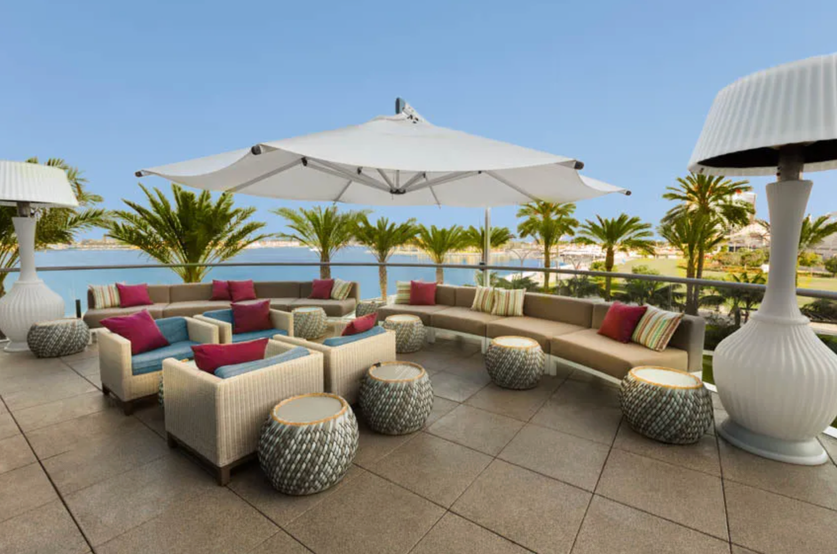 Outdoor patio with a large white umbrella, tan sofas, matching chairs with magenta pillows, and decorative floor lamps.