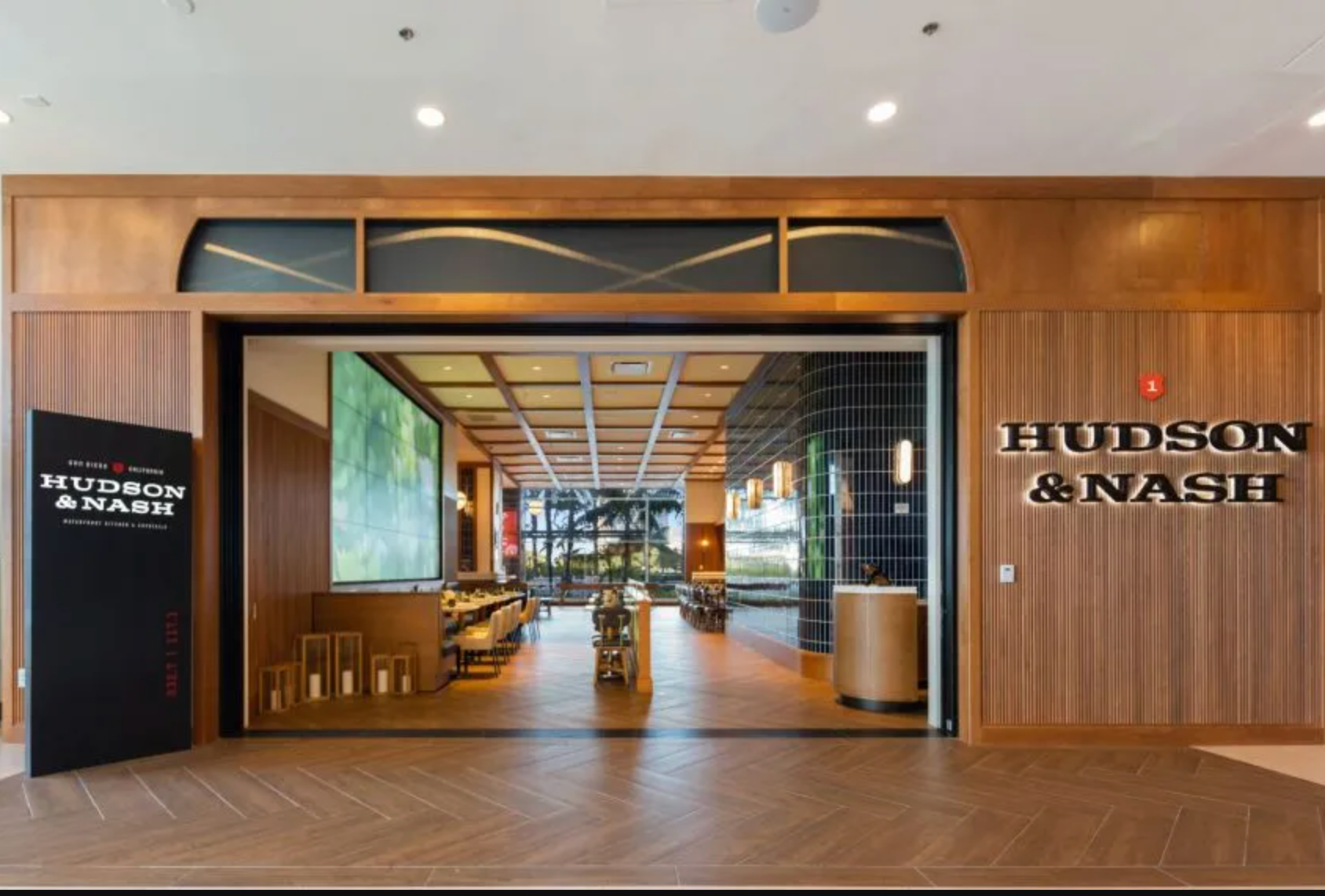 Entrance to Hudson & Nash restaurant with wooden paneling, large glass doors, and herringbone floor pattern.
