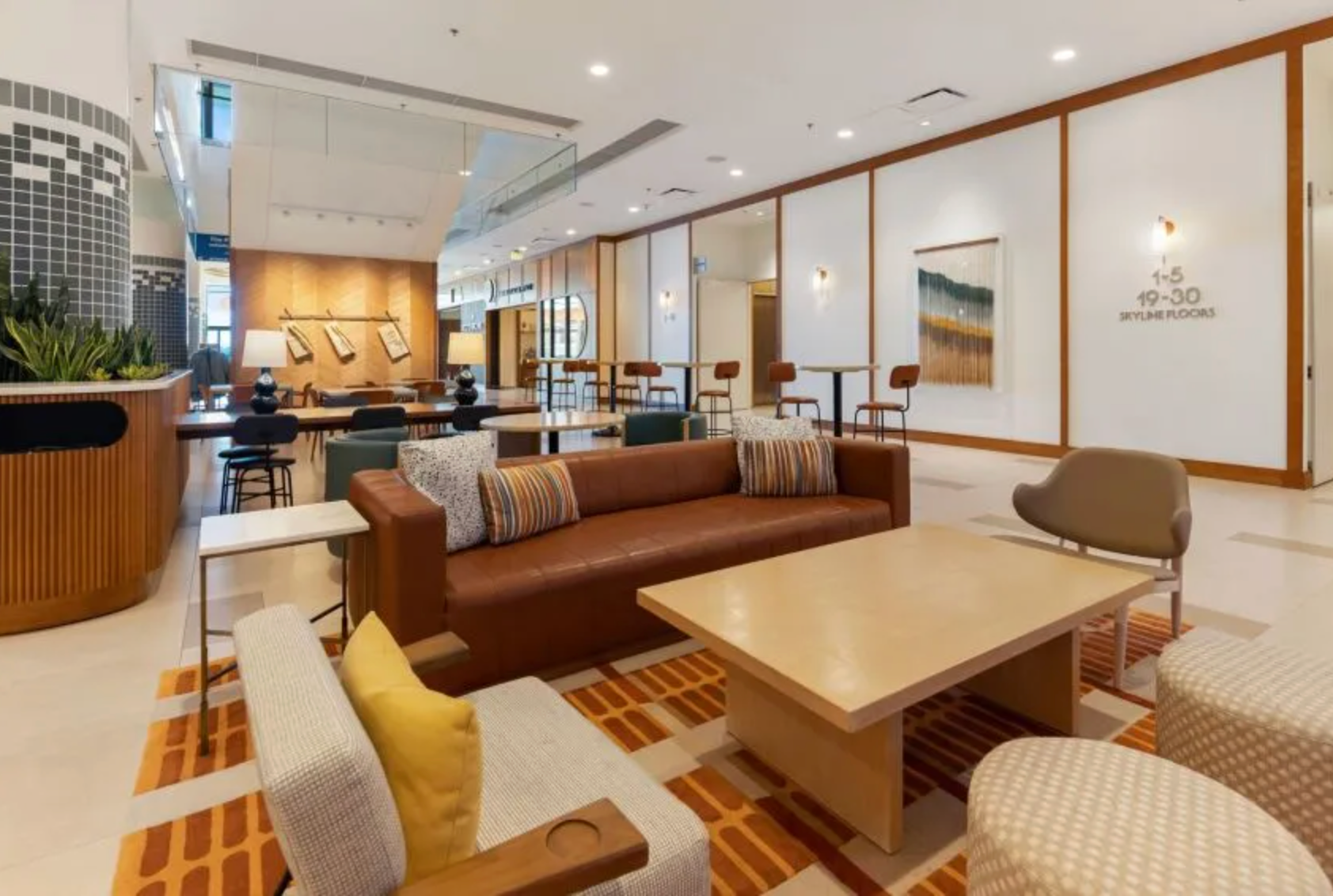 A modern, airy hotel lobby with comfortable brown leather sofas, armchairs, a coffee table, and an orange patterned rug.
