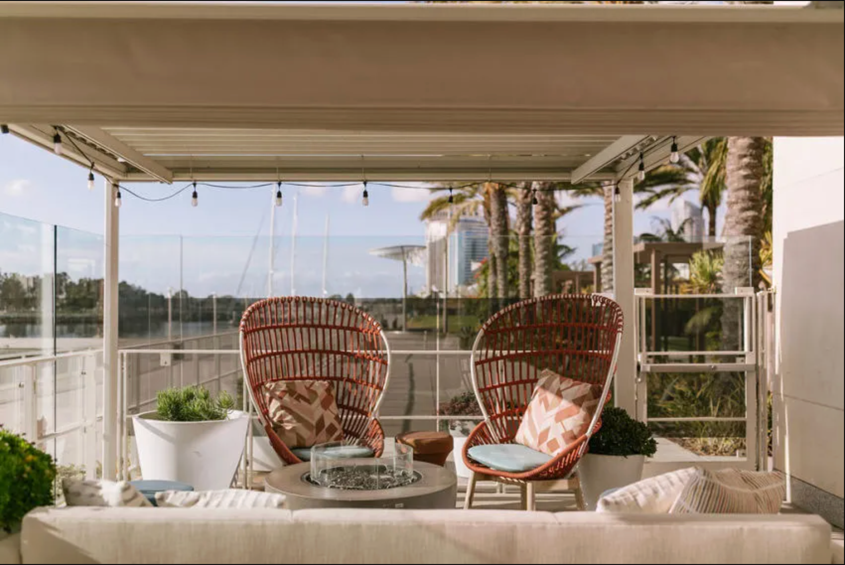 Patio overlooking a river with two wicker egg chairs, a central fire pit, and a cream sofa under a covered pergola.