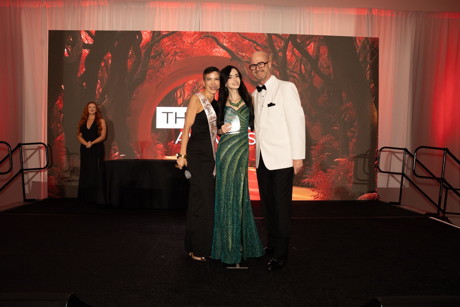 Three people pose for a photo on a stage at an awards event with a forest backdrop; the center person holds an award.