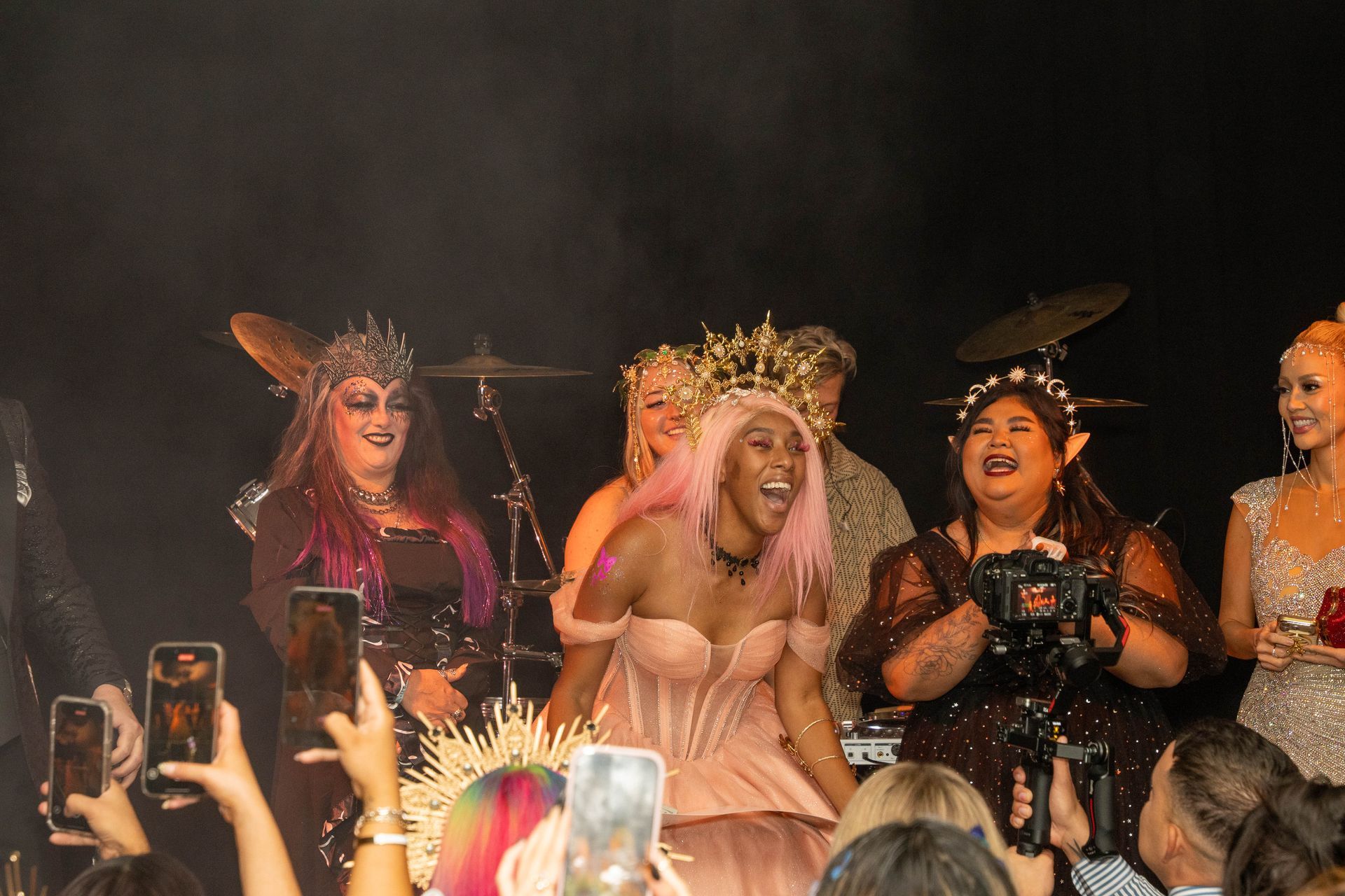 A joyful group of performers in ornate, fantastical costumes and headpieces stand on stage, smiling for a camera.