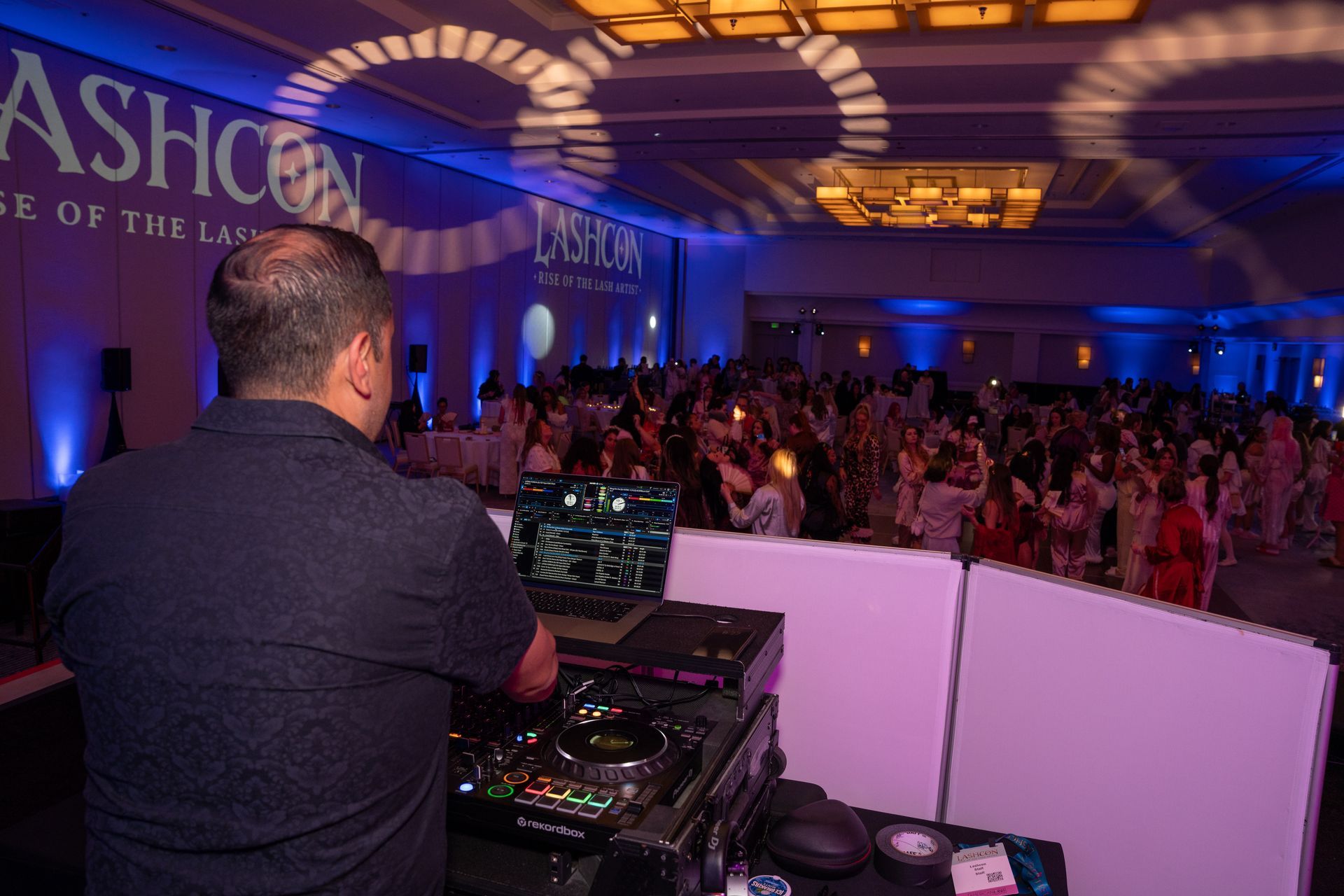 A DJ stands at a console with a laptop, looking out at a large crowd dancing in a brightly lit, decorated ballroom.
