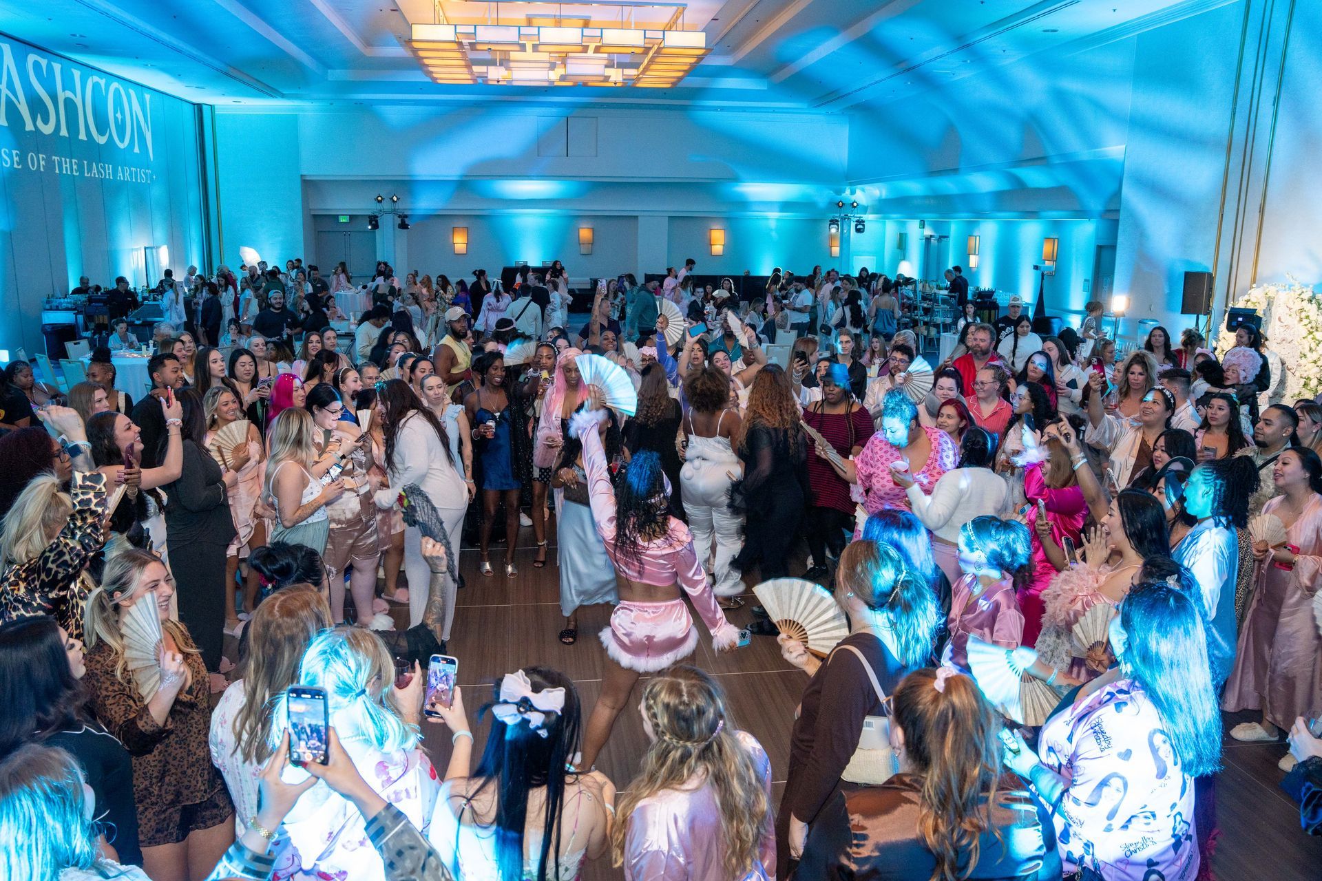 A large, energetic crowd dances in a brightly lit ballroom with blue ambient lighting and a high ceiling.