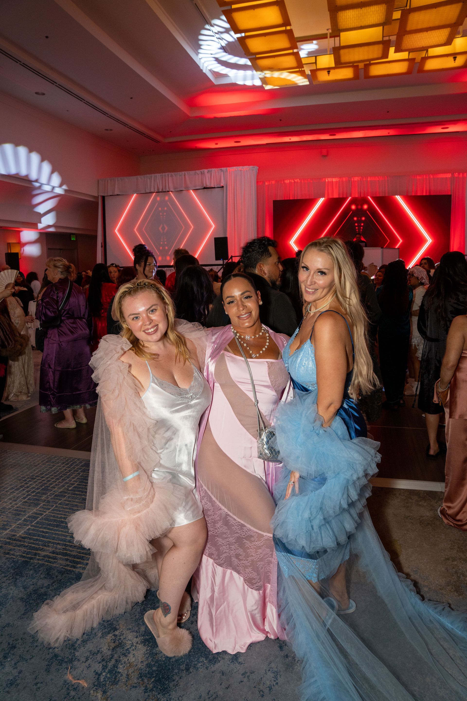 Three people in formal, tulle-detailed dresses—blush, pink, and light blue—pose at a brightly lit, festive indoor event.