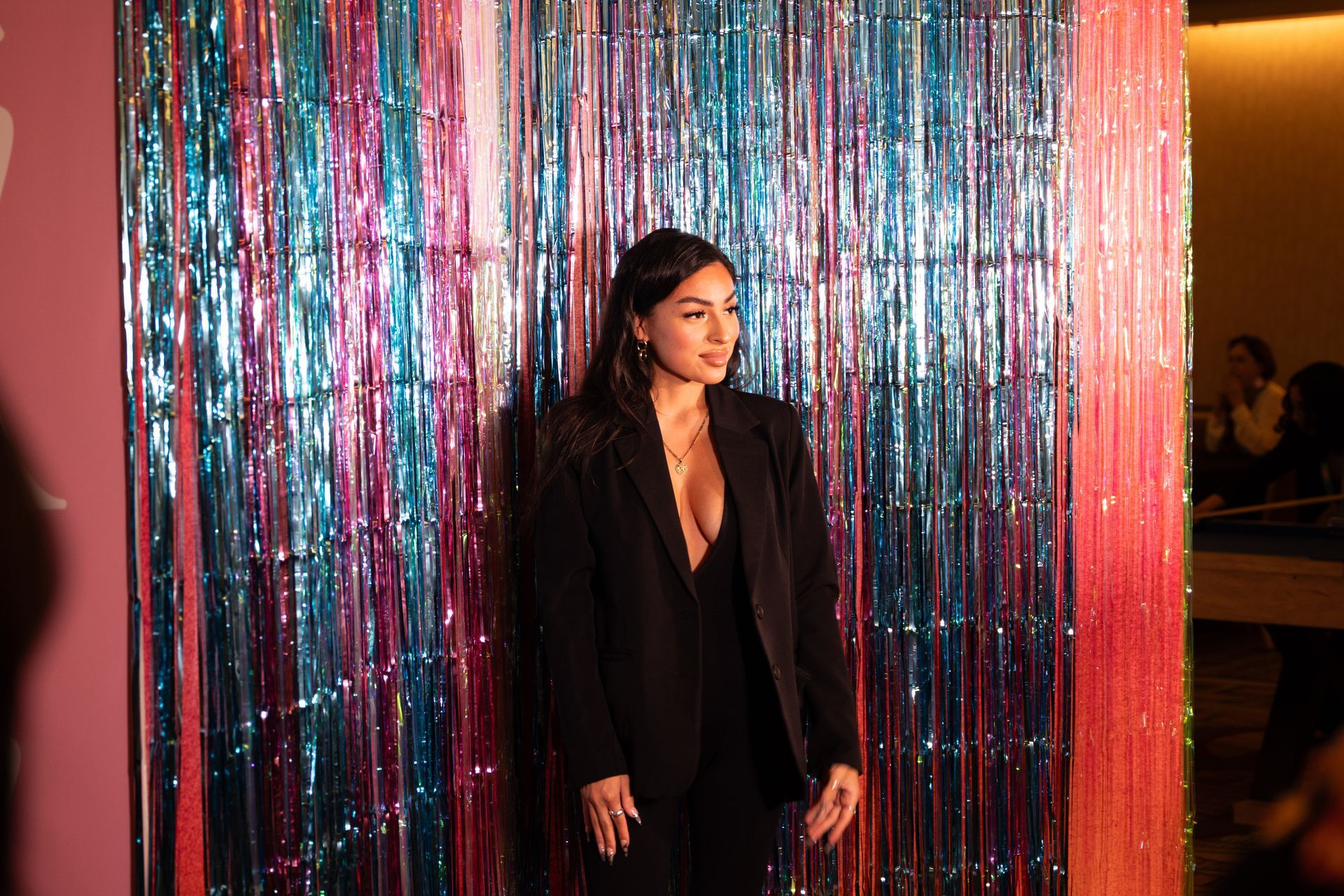 A woman in a black suit is standing in front of a colorful tinsel wall.