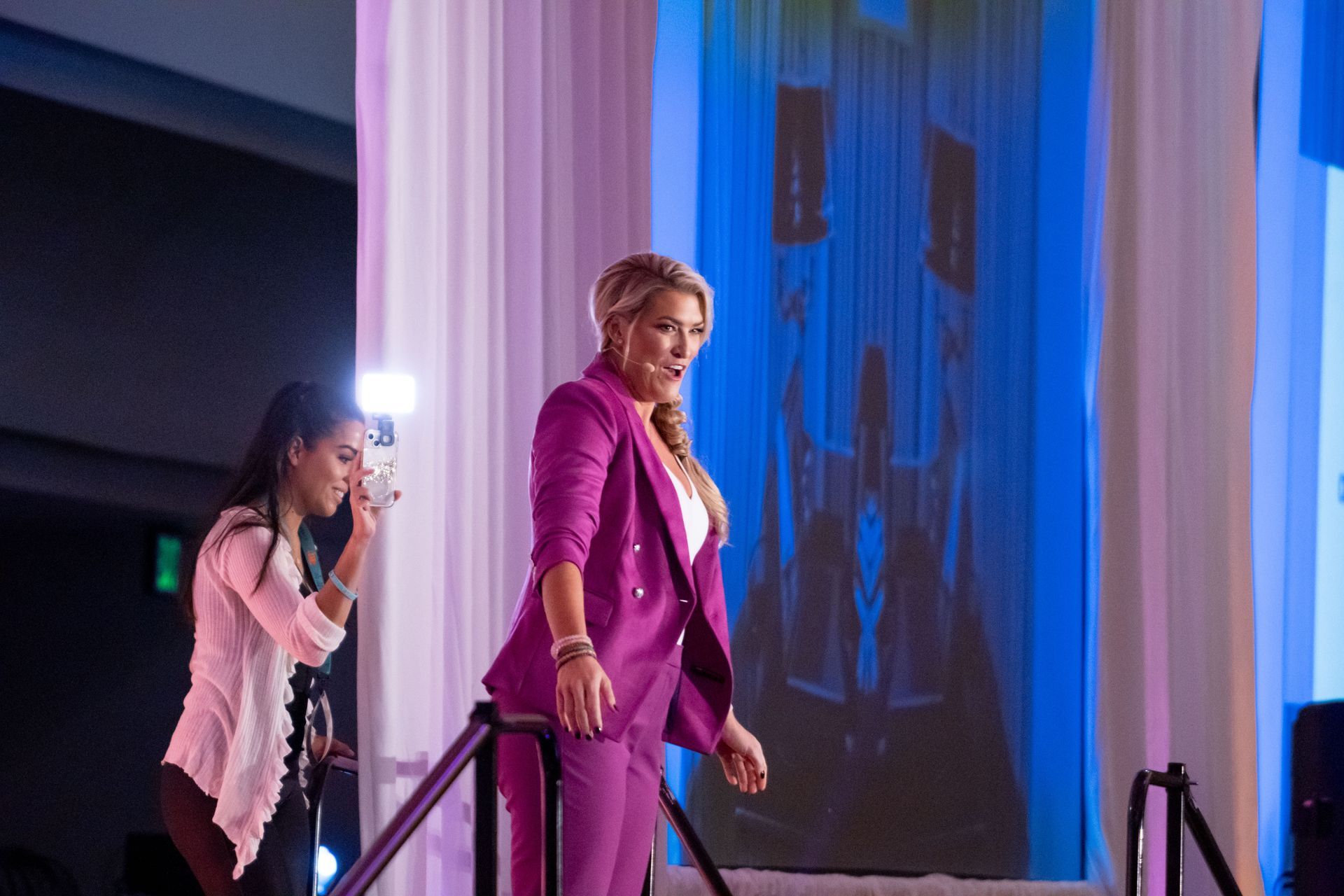 A woman in a purple suit is standing on a stage while another woman takes a picture of her.