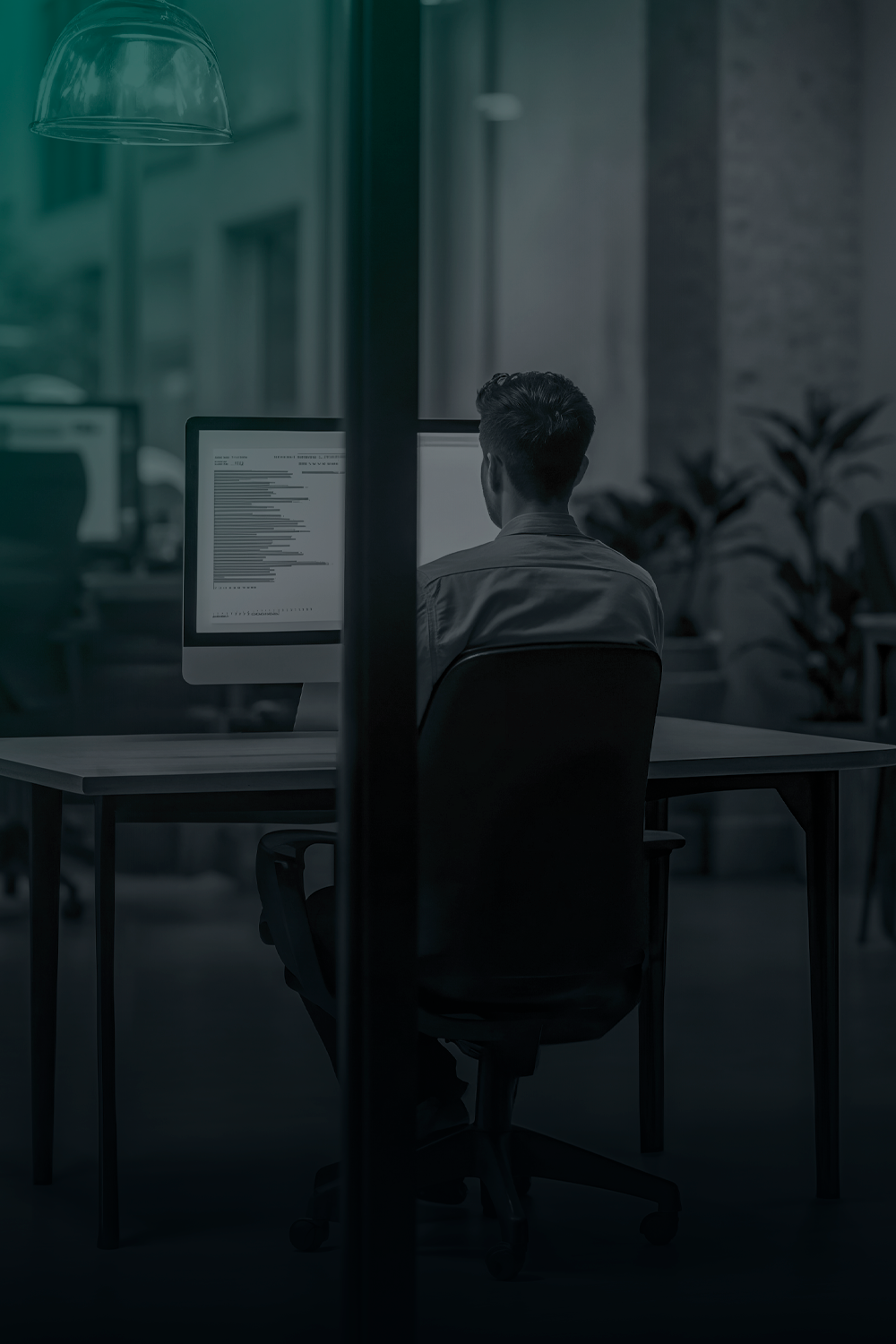 Pessoa sentada em frente a um computador, trabalhando em um escritório com pouca luz.