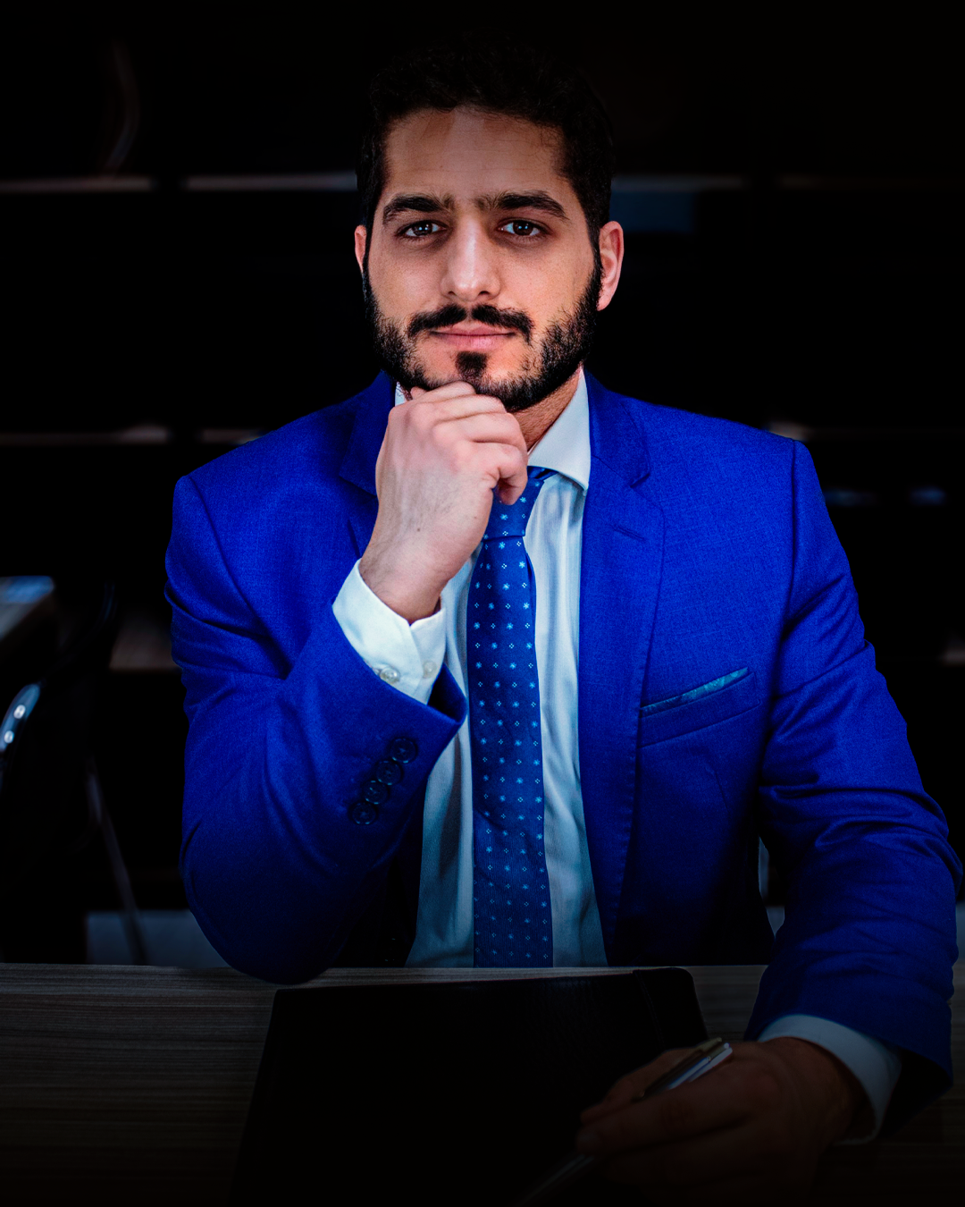 Homem de terno azul, mão no queixo, olhando para a câmera. Ambiente escuro.