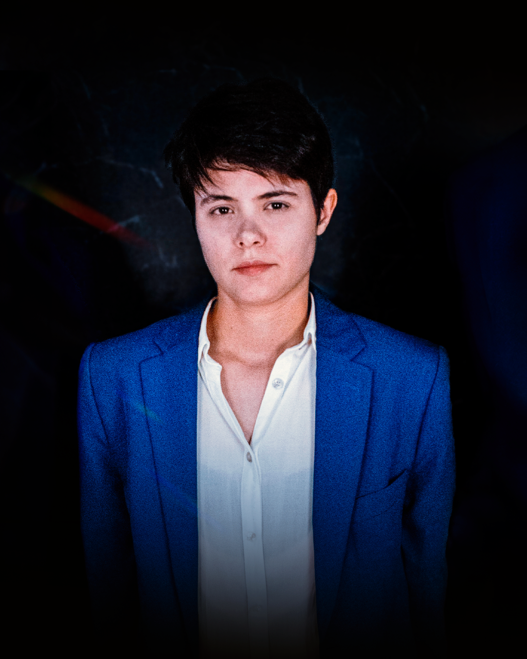 Mulher de blazer azul e camisa branca, olhando para o espectador. Fundo escuro com um prisma em arco-íris.