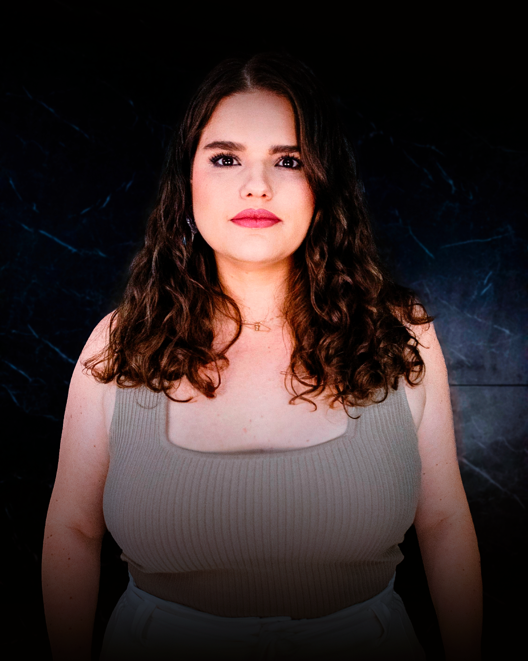 Mulher com blusa bege, cabelos castanhos cacheados, olhando para a câmera, fundo escuro.