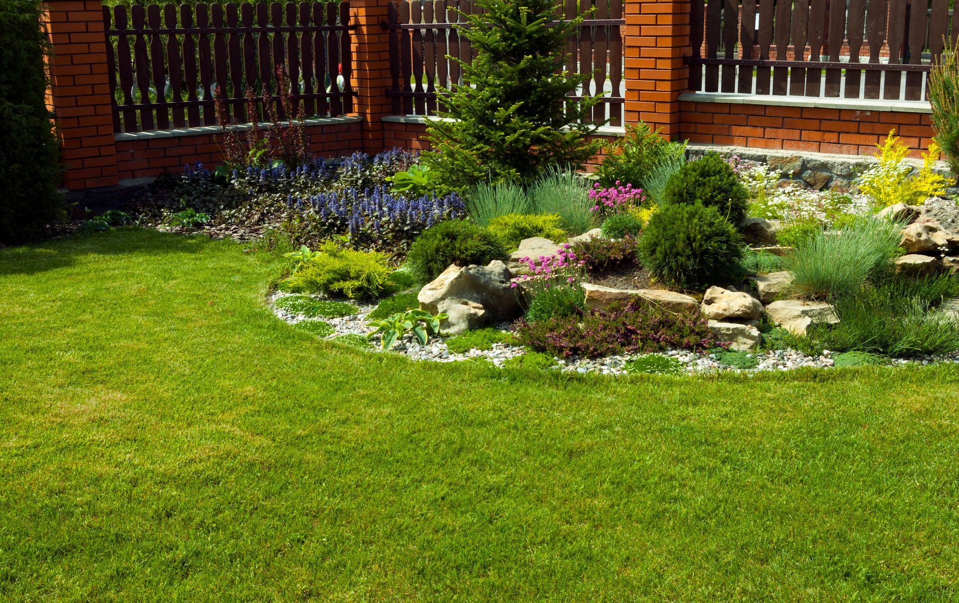 Small landscaped garden with green lawn, flowers, rocks, and brick fencing in the background