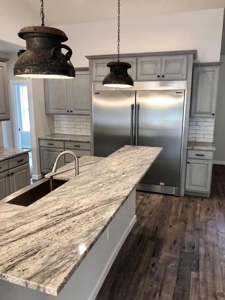 Kitchen with gray cabinets, stainless steel refrigerator, granite island, and hanging pendant lights.