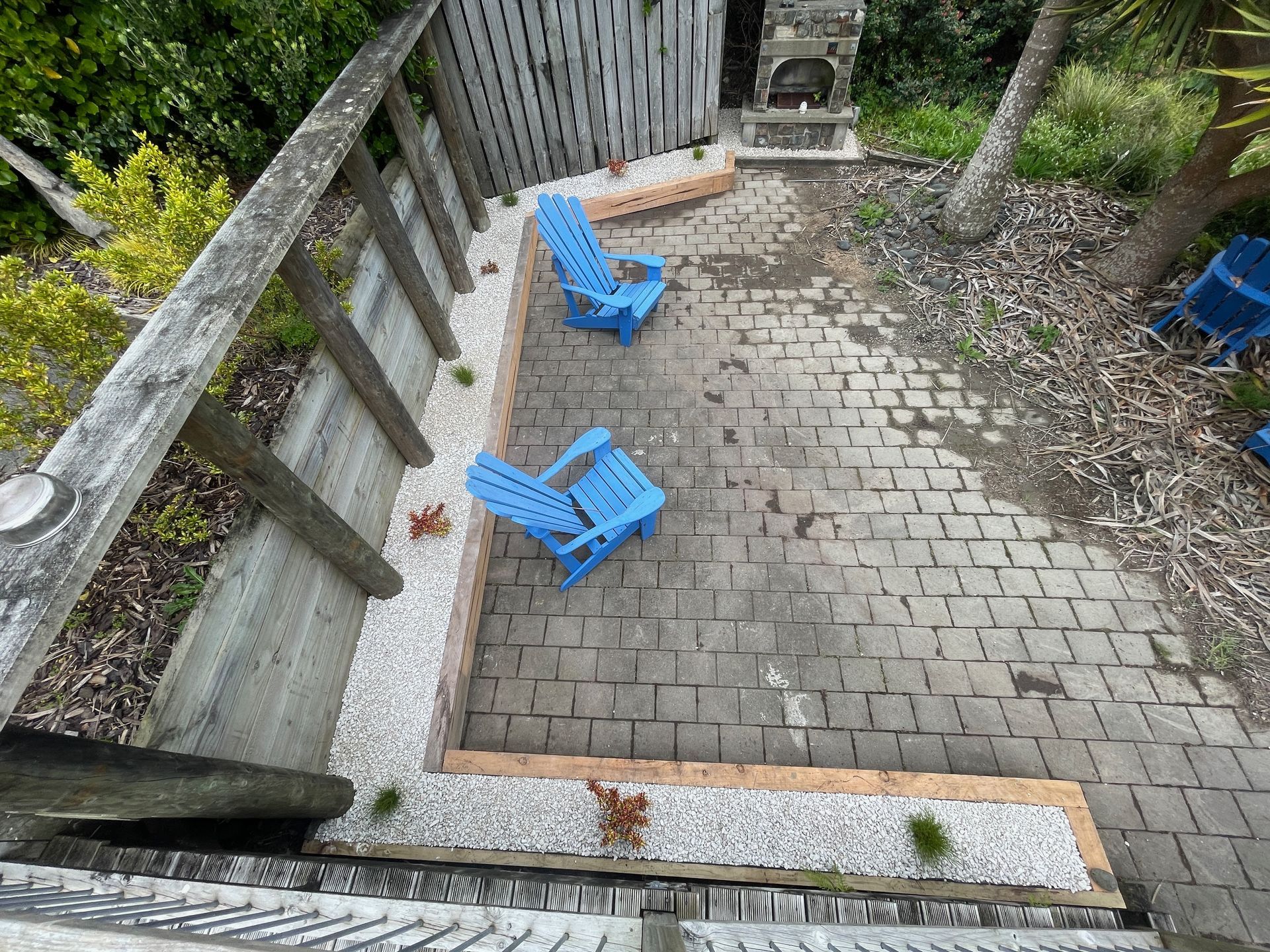 Two blue chairs on a brick patio surrounded by white pebbles and a wooden fence.
