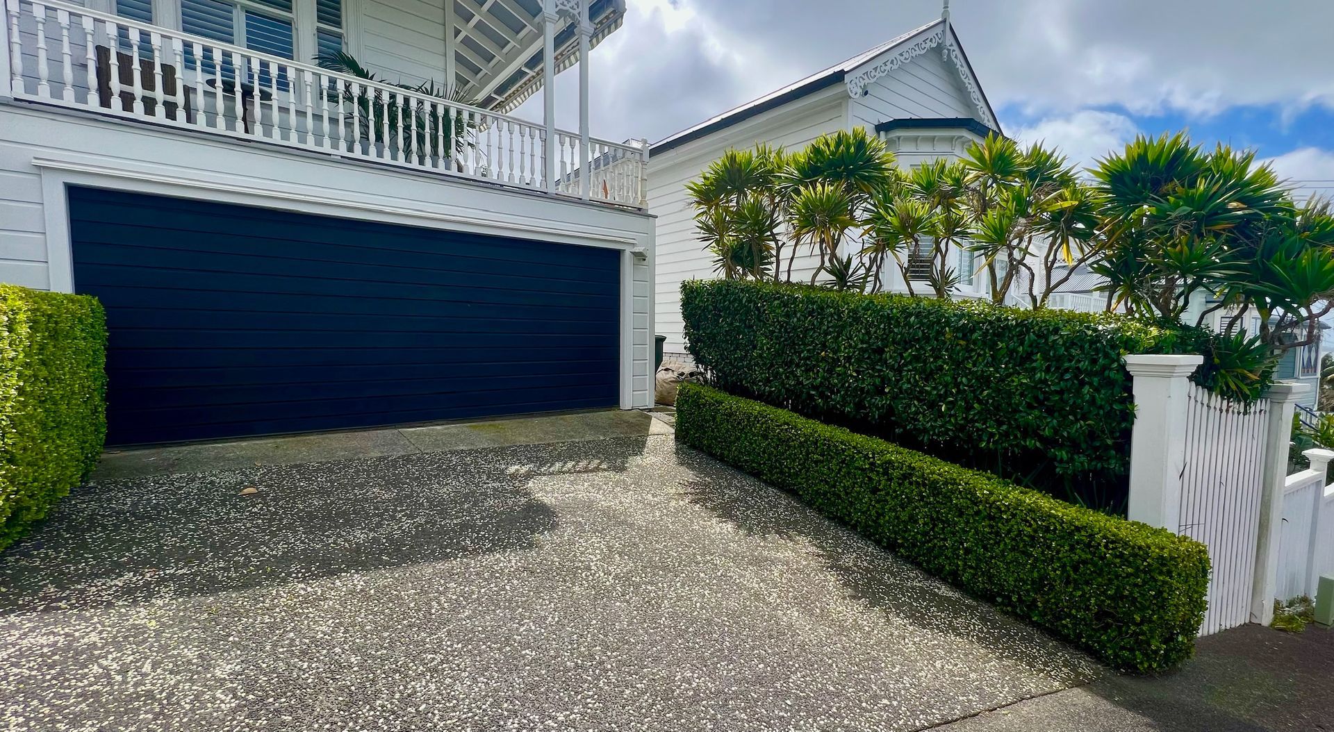 White house with black garage door; driveway lined with hedges, cloudy sky.