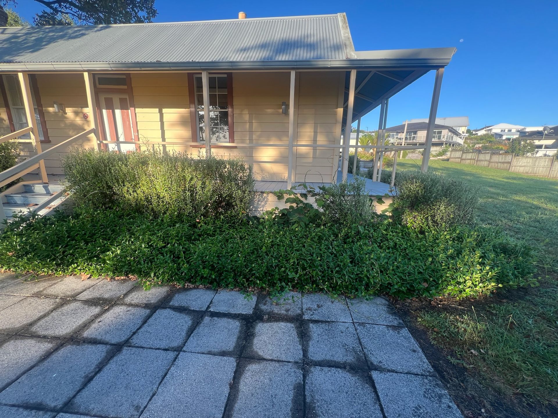 Yellow building with a porch, steps, and green bushes in front, with a stone walkway and blue sky.