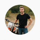 Man in black shirt and jeans standing next to an orange and white dirt bike.