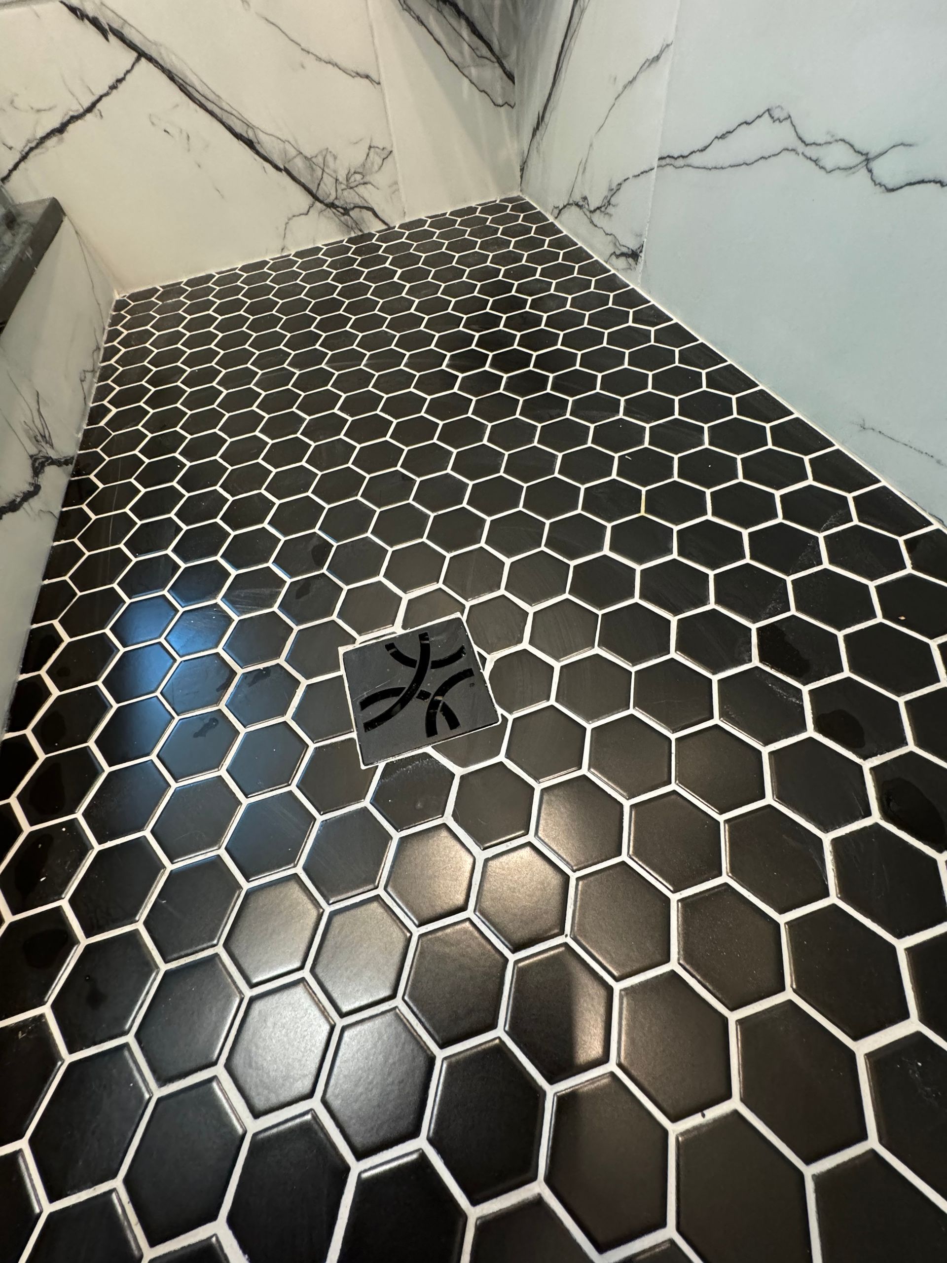 A shower stall with black and white hexagon tiles and a drain.
