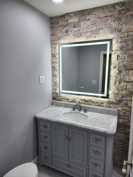 Bathroom with a grey vanity, a lit mirror on a brick accent wall, and a toilet.