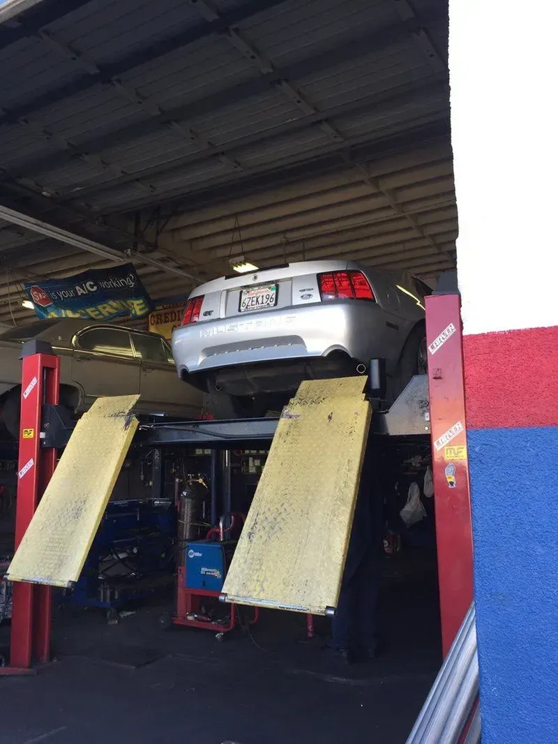 Silver car raised on a lift inside a garage, showing undercarriage. Yellow ramps, red lift columns.