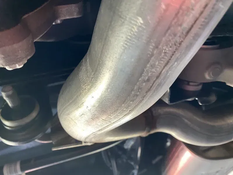 Undercarriage view of a curved exhaust pipe. The pipe is silver, welded together, and visible beneath the vehicle.