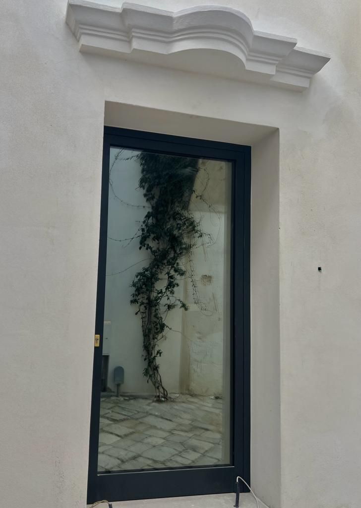 Finestra con cornice scura che riflette il cortile e la pianta rampicante sul muro bianco.