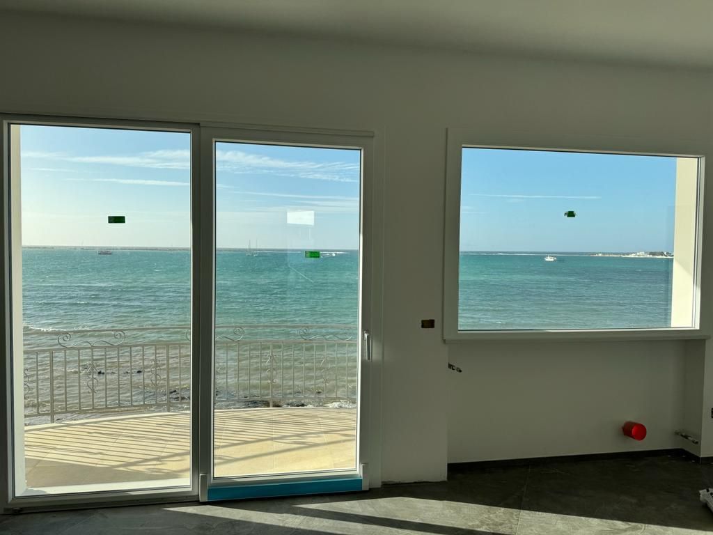 Vista interna di una stanza con vista sull'oceano attraverso porte e finestre in vetro. Acqua azzurra, cielo luminoso.