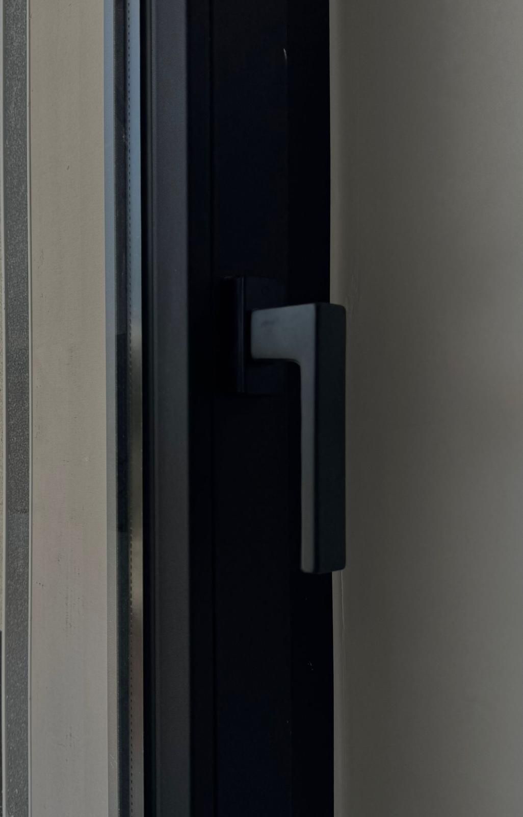 Maniglia nera per finestra su telaio scuro, con parete beige chiaro sullo sfondo.