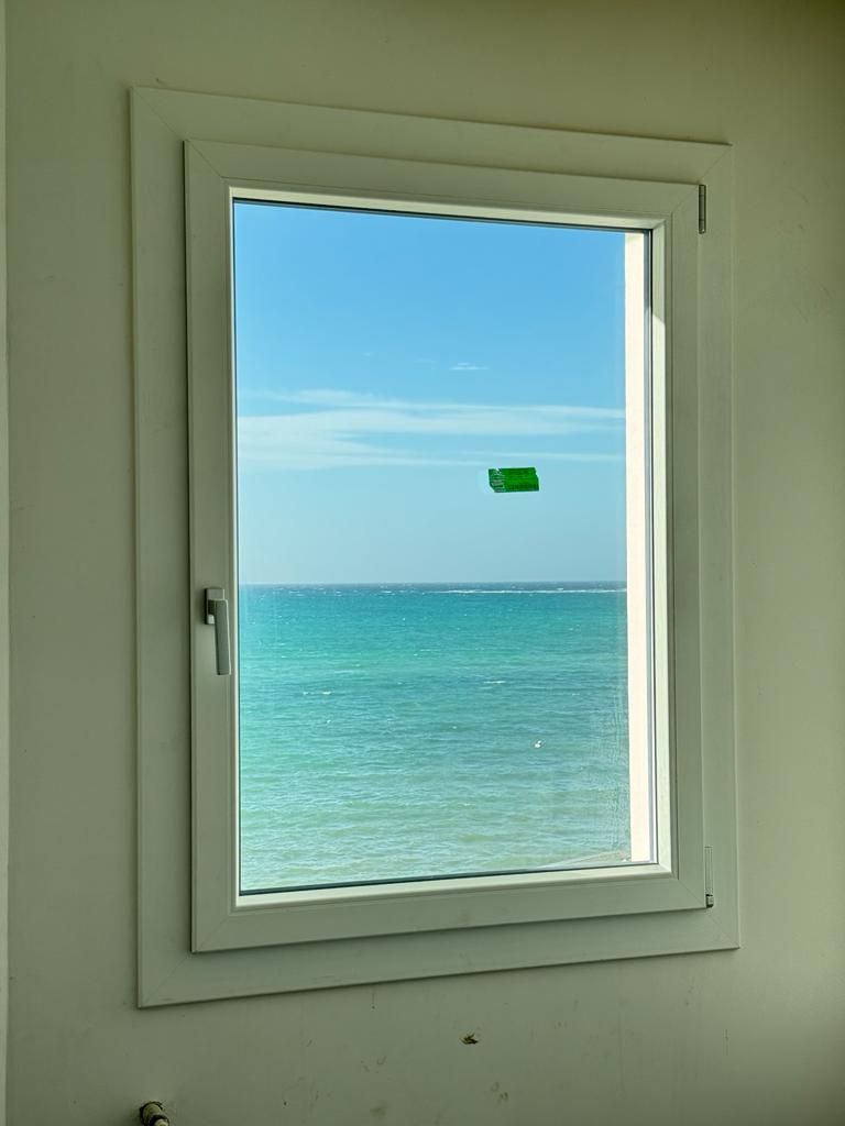 Finestra con vista sull'oceano turchese e sul cielo azzurro. Un quadrato verde si intravede nel cielo.