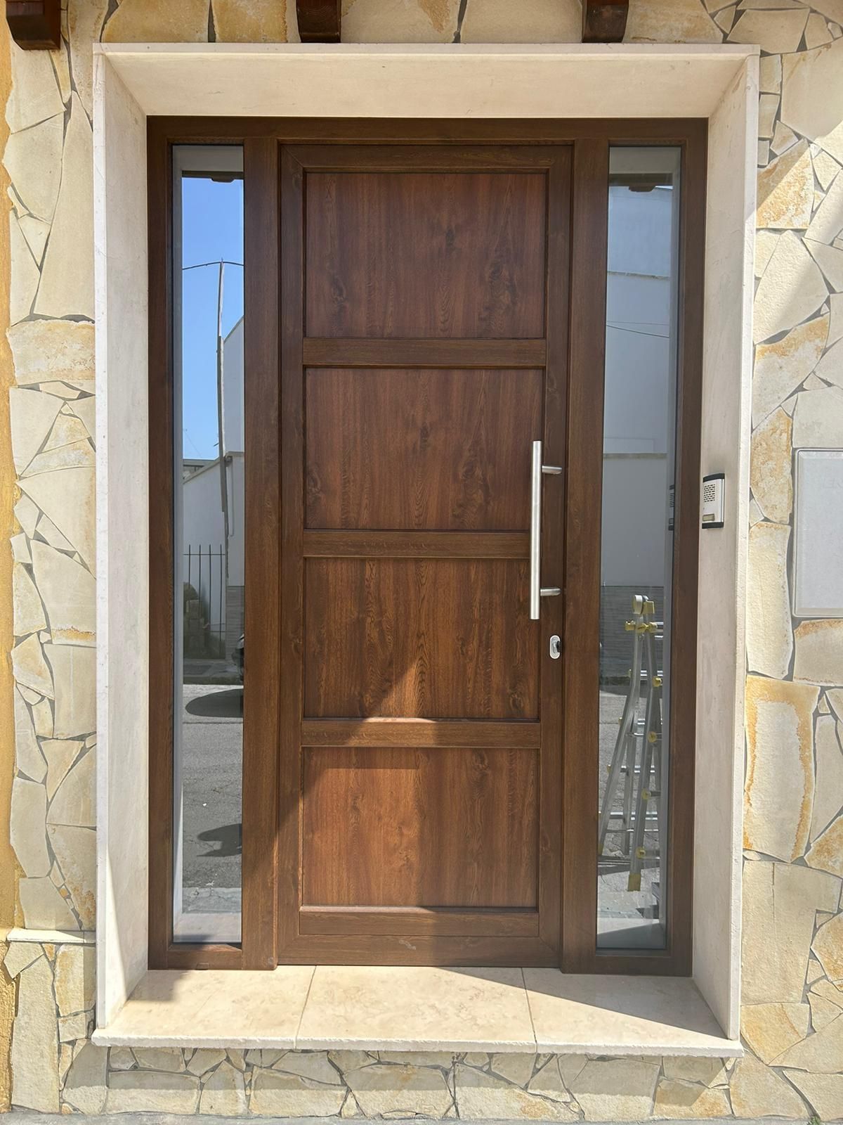 Porta d'ingresso marrone con vetri laterali. Incastonata in una cornice in pietra, con maniglia in metallo.