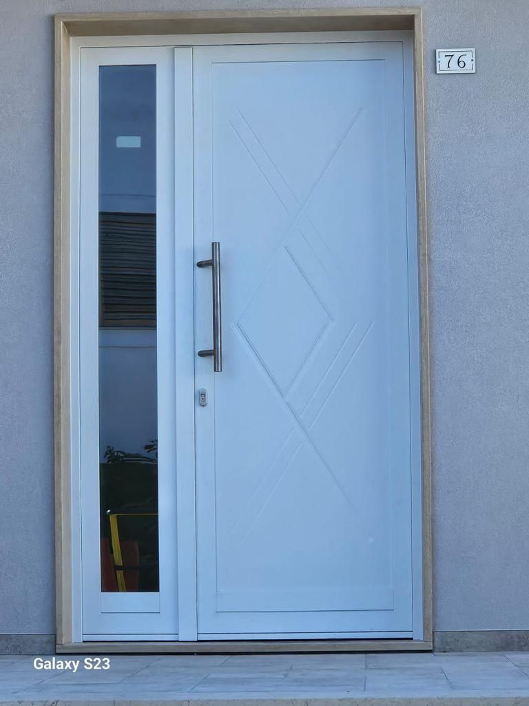 Porta d'ingresso bianca con finestra laterale. Maniglia argentata. Facciata esterna dell'edificio.