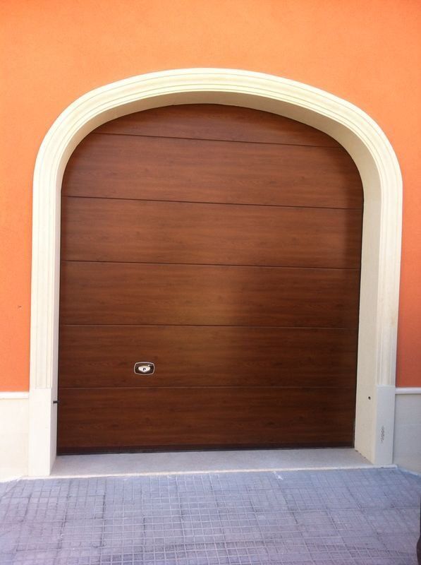 Porta da garage marrone inserita in una cornice ad arco con finiture bianche, contro una parete arancione.