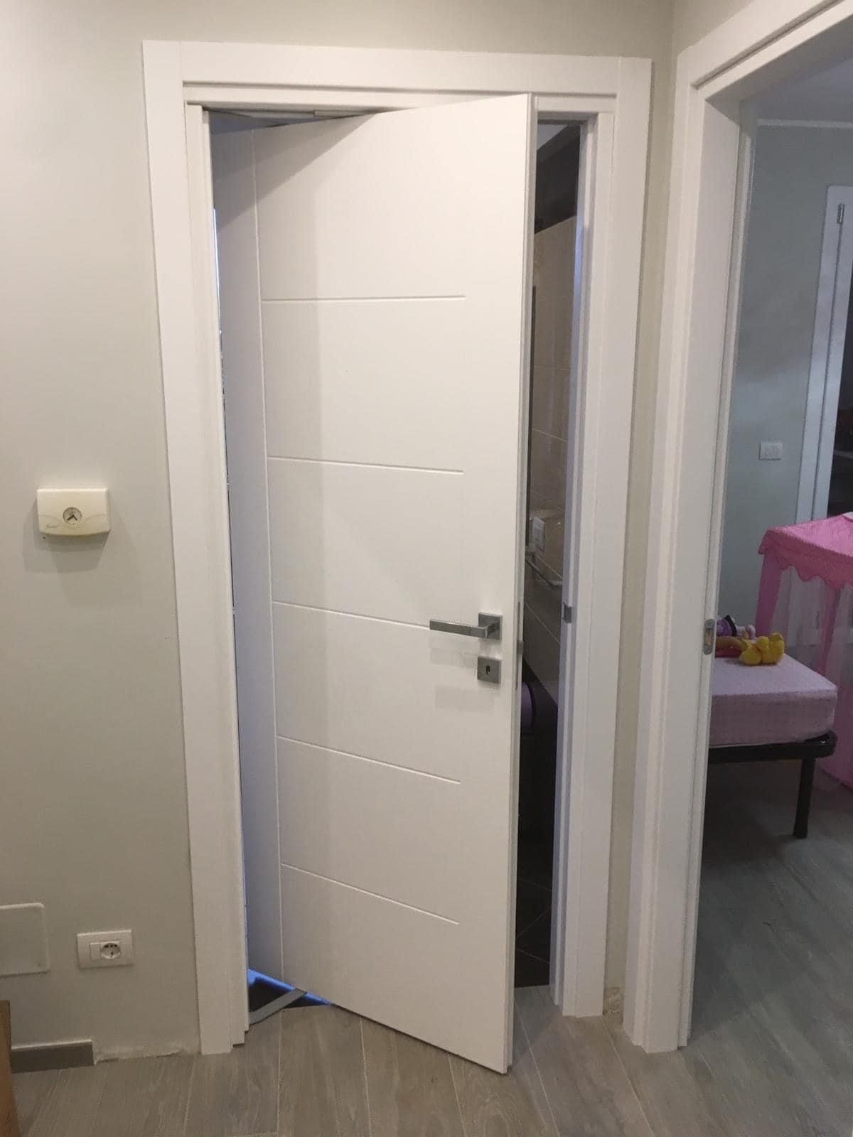 Una porta bianca leggermente aperta nel corridoio rivela un interno buio, con una stanza visibile sulla destra.