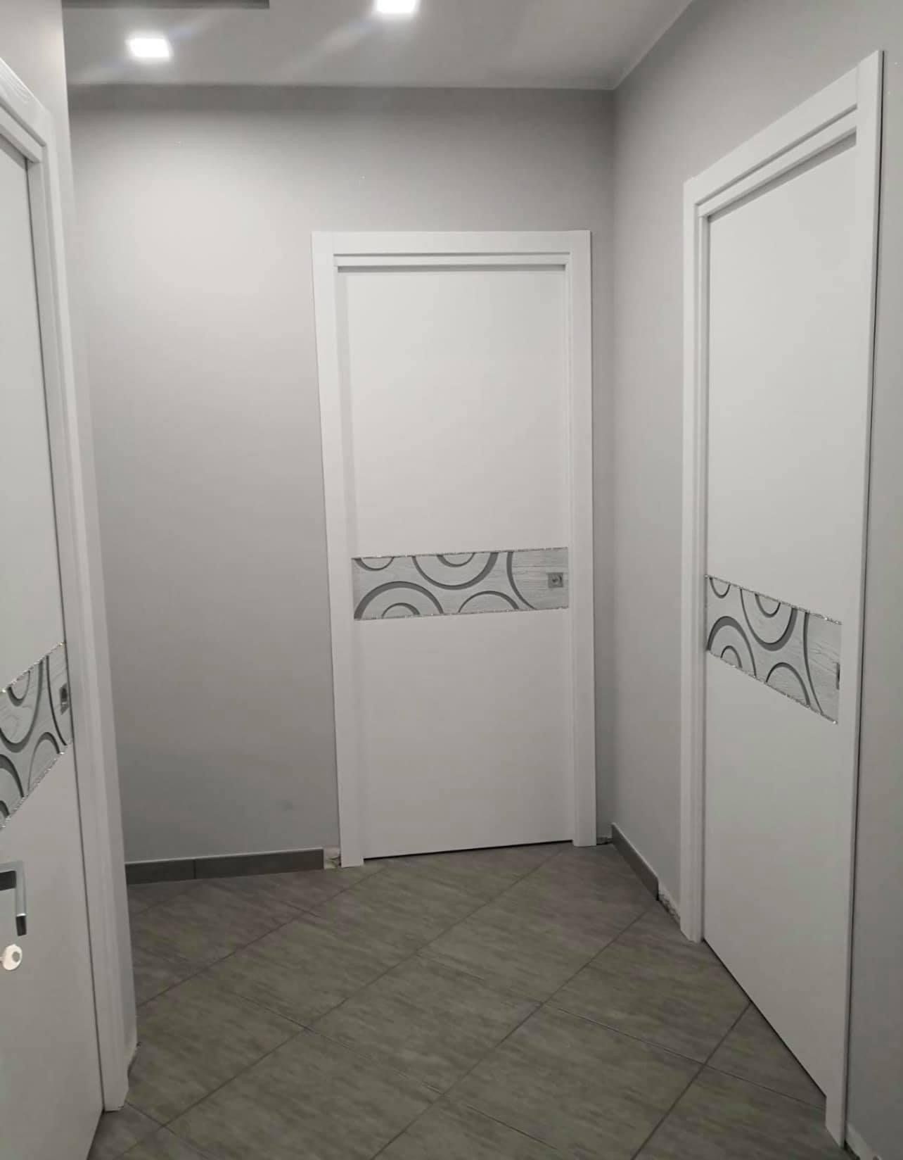 Corridoio con tre porte bianche, ciascuna con un pannello decorativo in vetro e pavimento piastrellato.