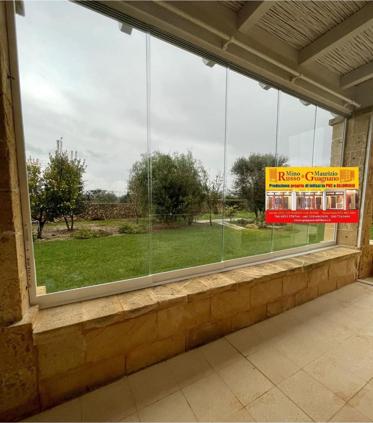 Vista di un giardino attraverso pannelli di vetro. Facciata in pietra beige, erba, alberi e un cartello giallo.