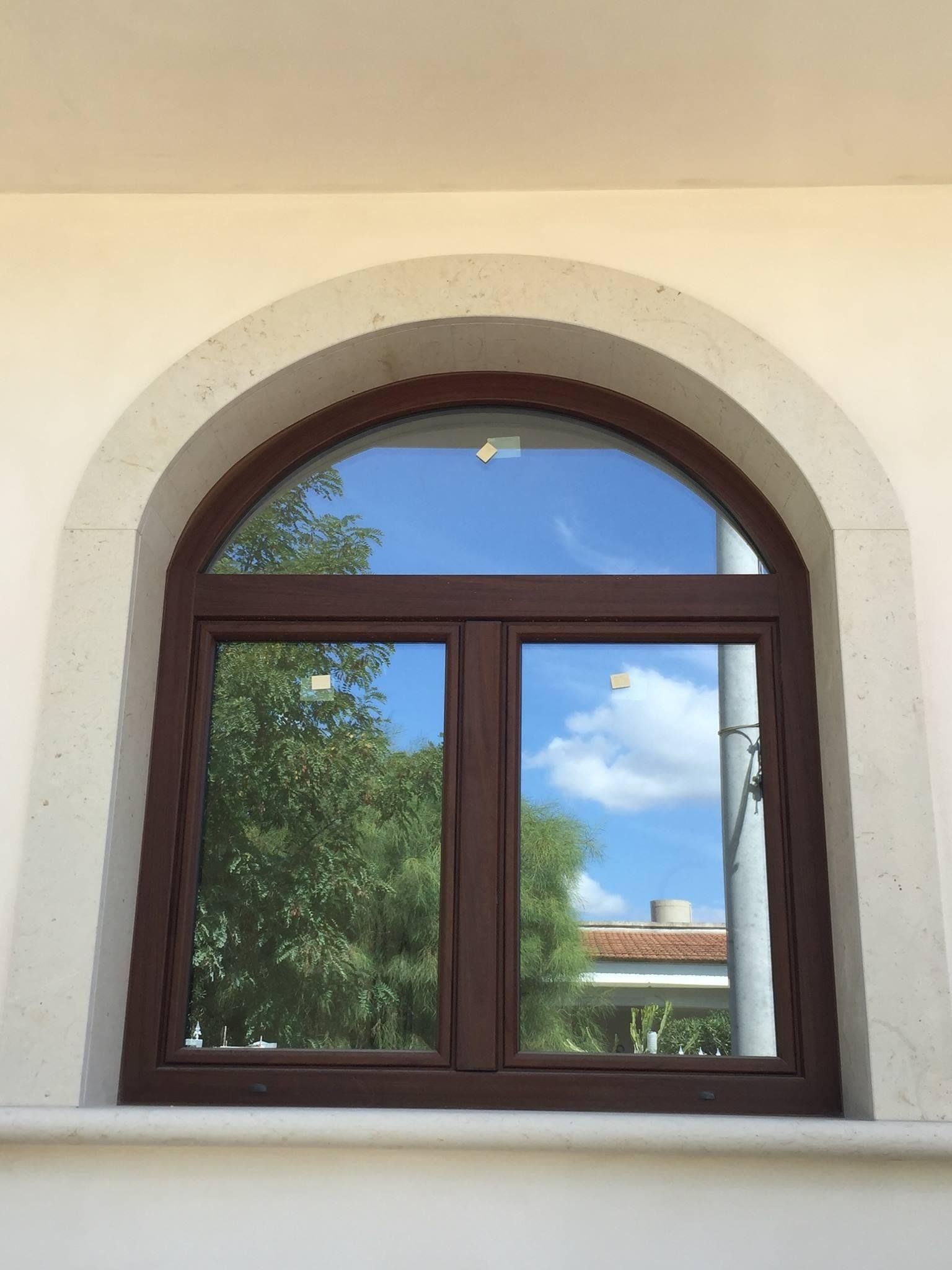 Finestra ad arco marrone che riflette il cielo e gli alberi, incastonata nel muro bianco.