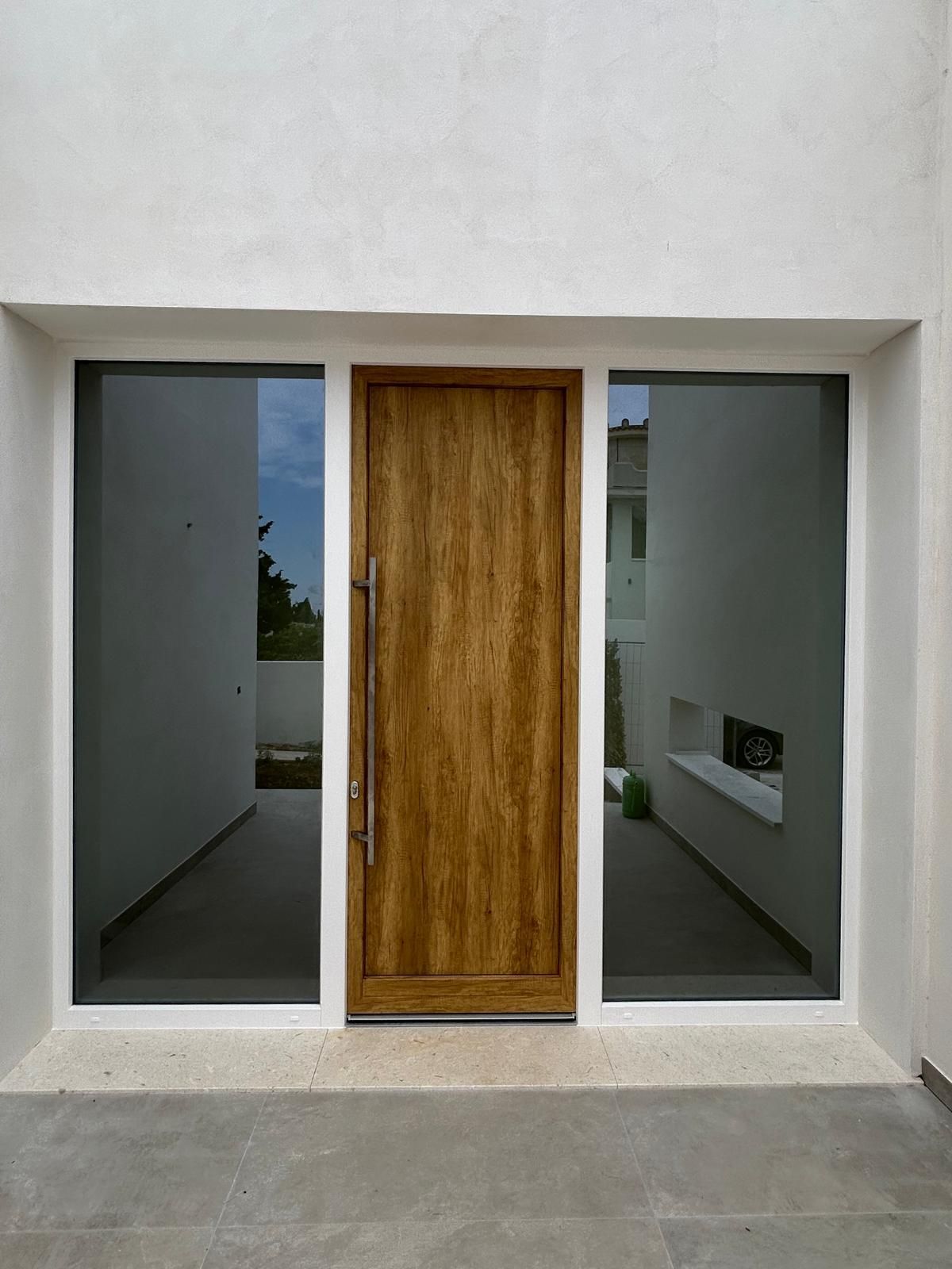 Porta in legno affiancata da due grandi finestre, inserita nella facciata bianca dell'edificio.