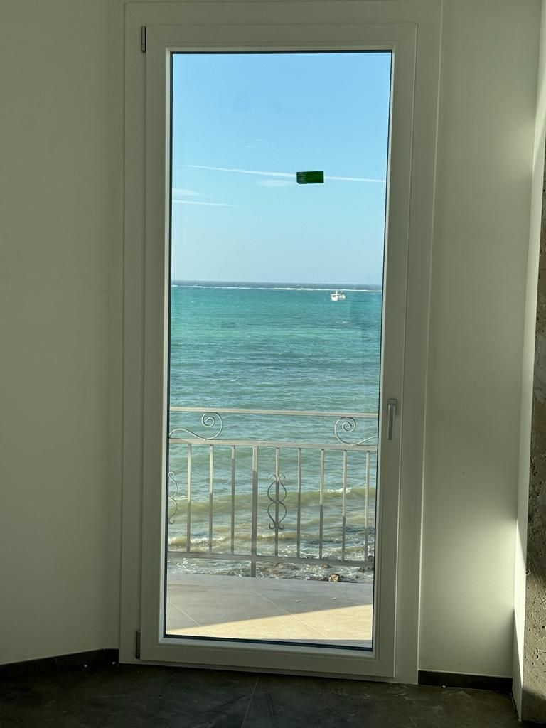 Porta con vista sull'oceano: acqua turchese, ringhiera bianca, cielo azzurro, nastro verde sulla finestra.