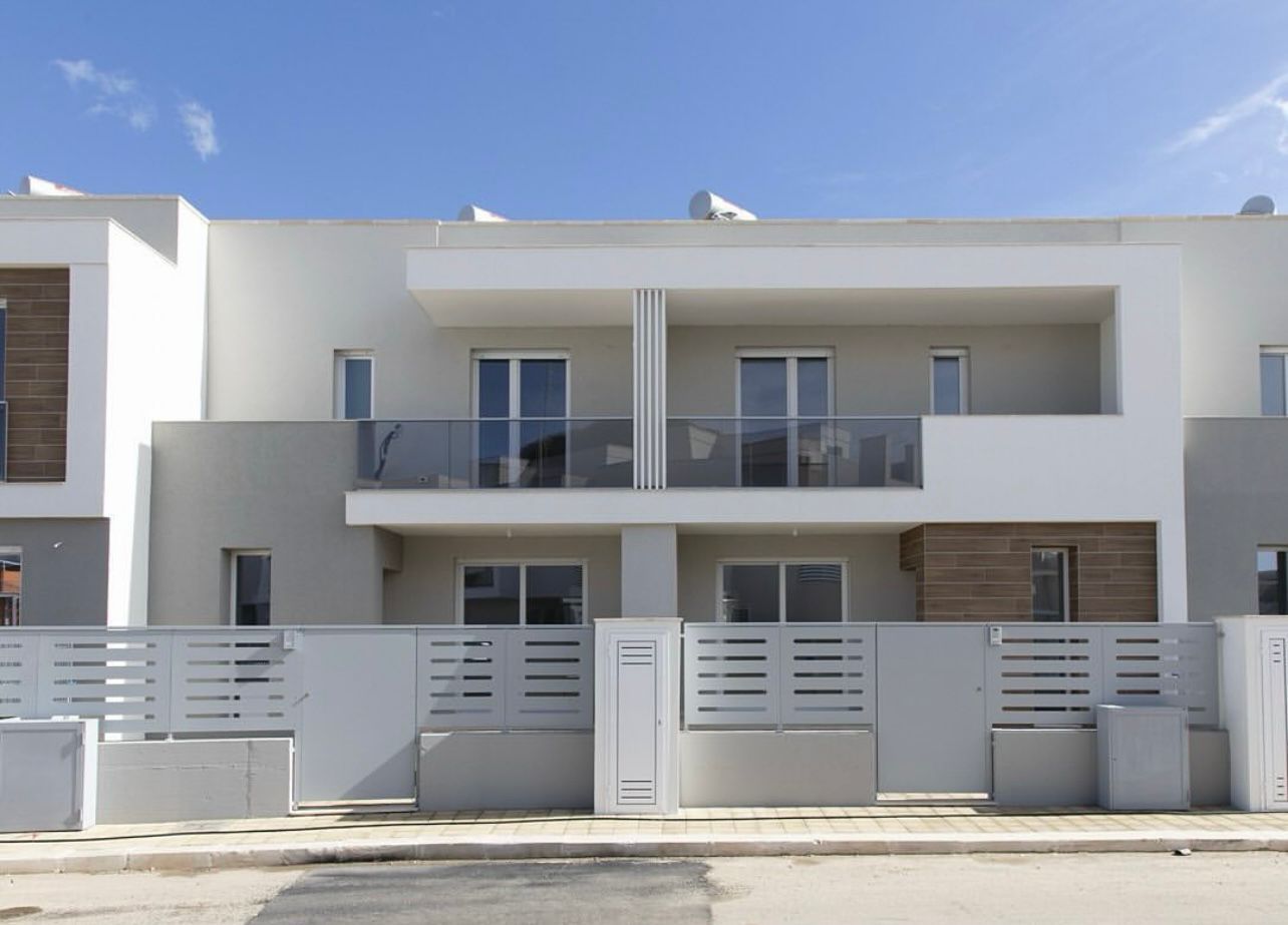 Moderne case a schiera bianche e grigie con balconi in vetro e una recinzione a doghe orizzontali sotto un cielo azzurro.