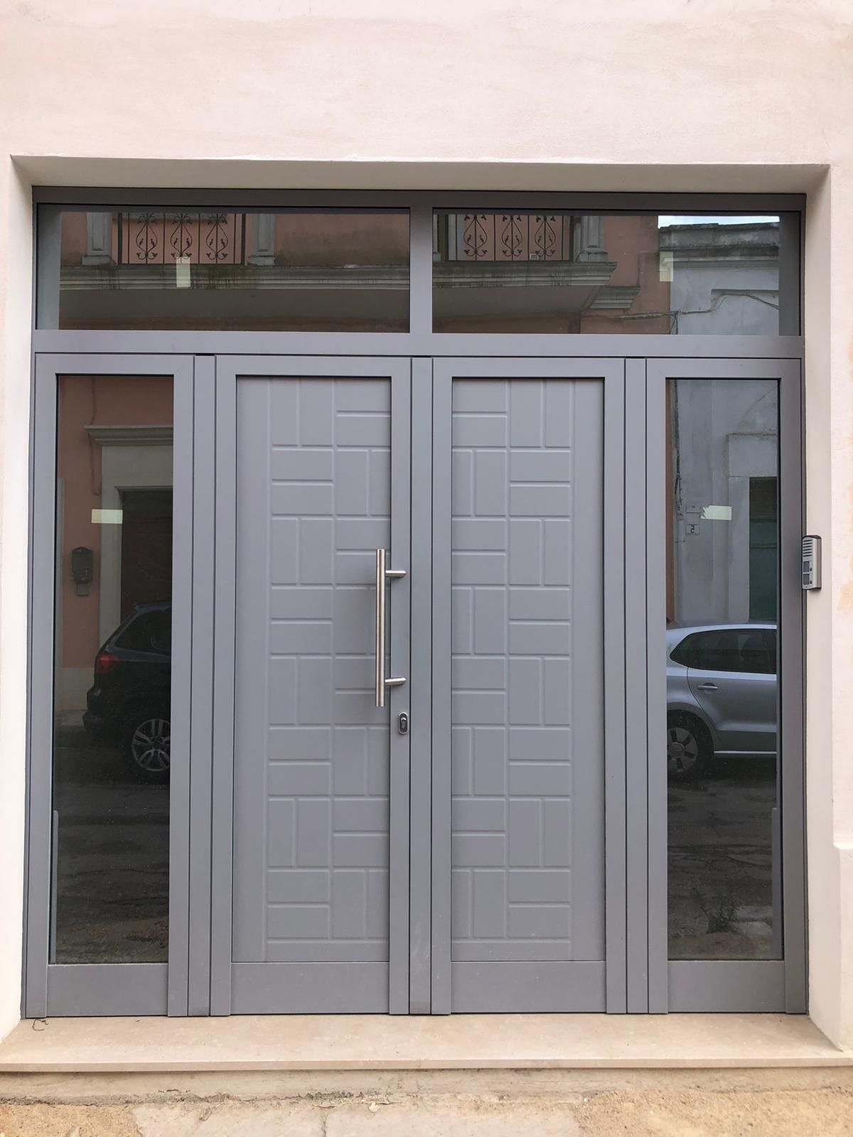 Doppie porte grigie con vetri laterali in vetro e finestra a traverso; ingresso di un edificio.
