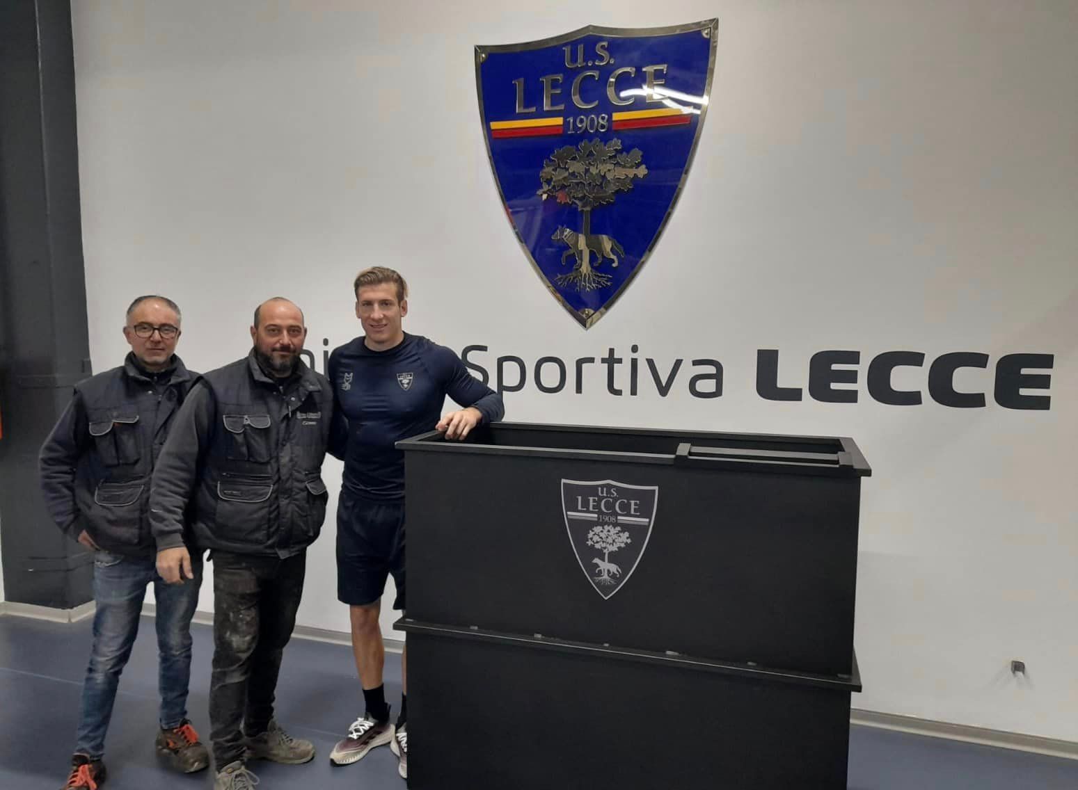 Tre uomini posano davanti al logo dell'US Lecce. Uno di loro è appoggiato a una scatola nera.
