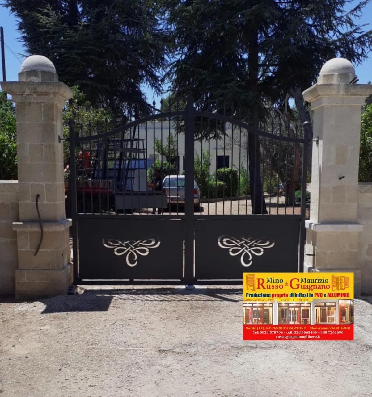 Cancello in ferro battuto nero con motivo decorativo tra pilastri in pietra, cartello pubblicitario sulla destra.